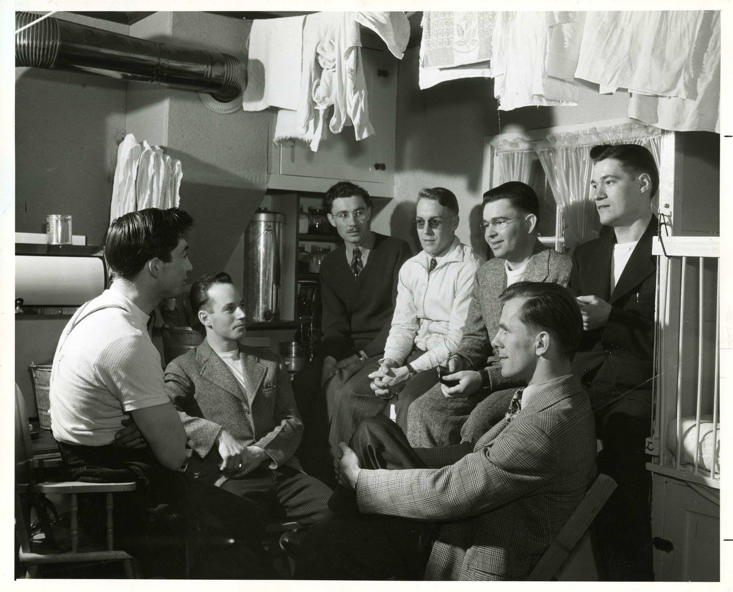 A photograph of seven men inside the kitchen of a home in Veterans' Village at the University of Manitoba Campus. Most, if not all, of the men are likely students. House is likely the home of the individual on the far left of the image. Photograph is dated as having been taken around 1948 by the National Film Board of Canada.<br />
Source: University Relations & Information Office fonds