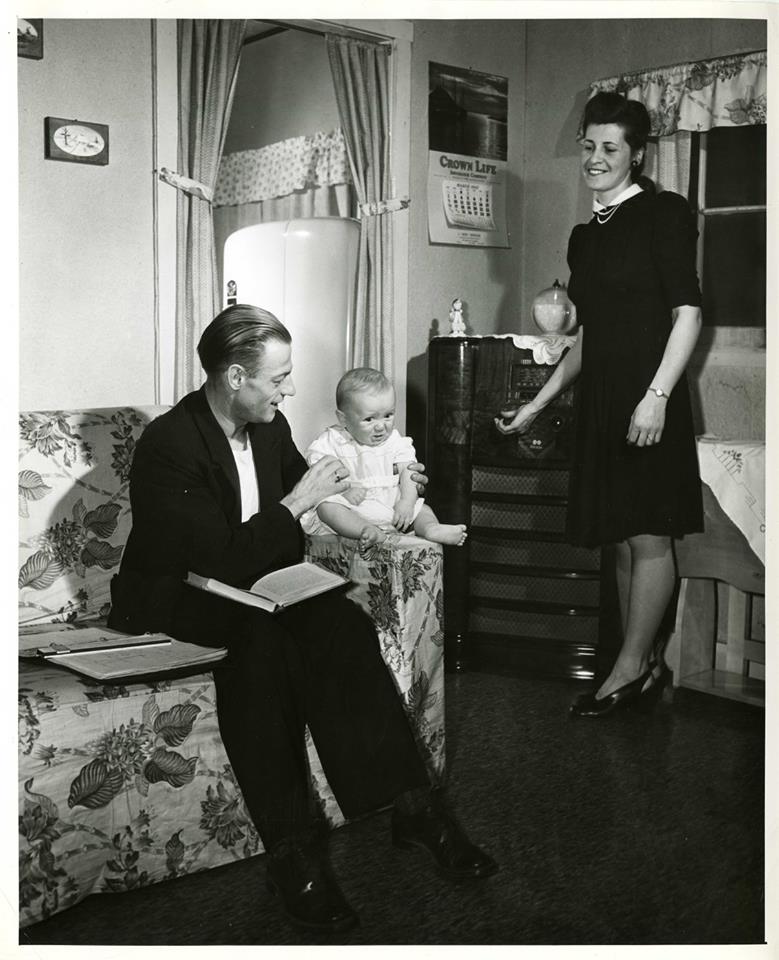 A photograph of a family inside the living room of a home in Veterans' Village at the University of Manitoba Campus. A man is seated on a couch, shown with books beside him and holding an infant. A woman appears to be adjusting the dial of a hi-fi player. Photograph is dated as having been taken around 1948 by the National Film Board of Canada.