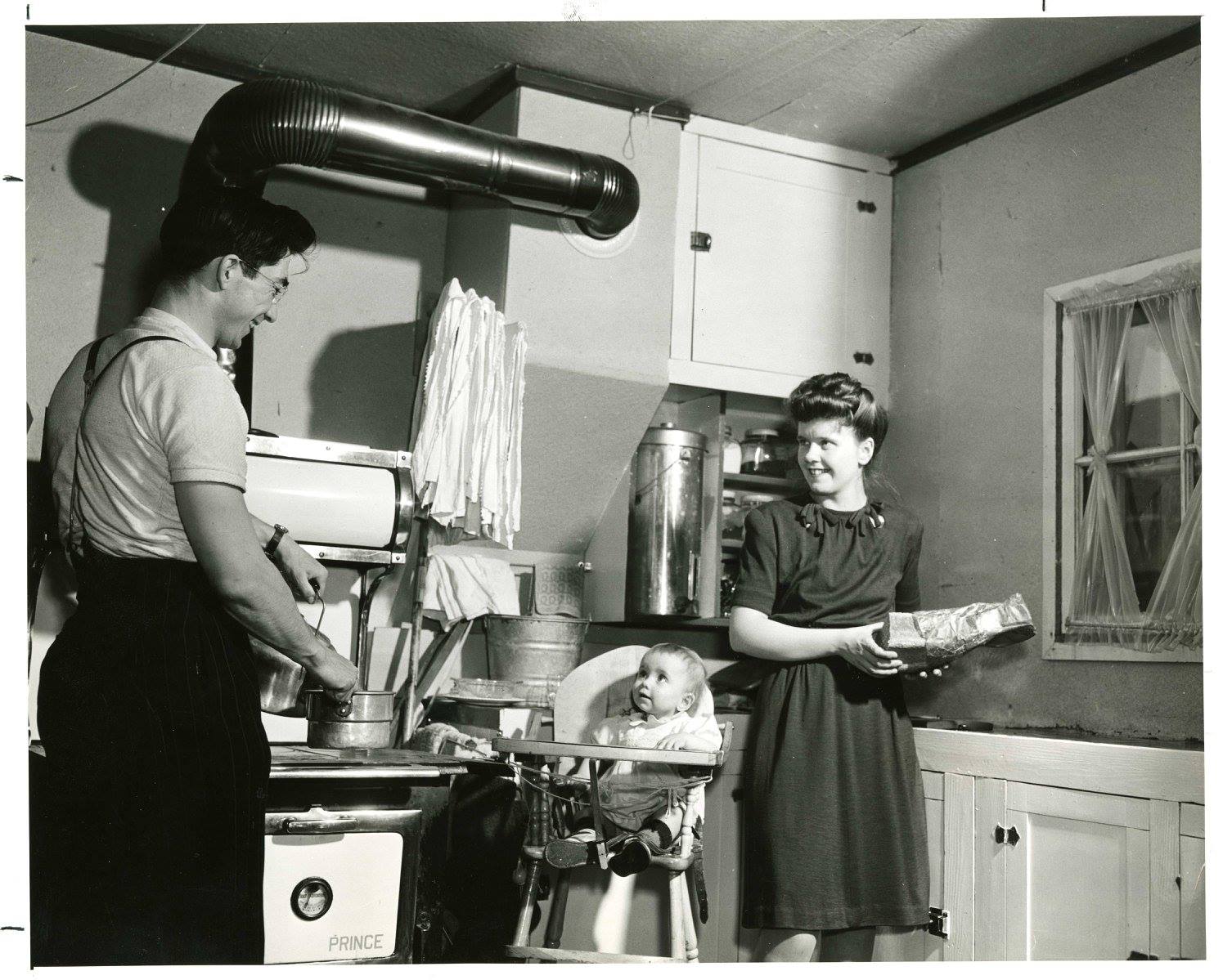 A photograph of a family inside the kitchen of a home in Veterans' Village at the University of Manitoba Campus. A man is standing at the stove with a pot, an infant is in a high chair, and a woman appears to be holding a loaf of bread and a bag in her hands. Photograph is dated as having been taken around 1948 by the National Film Board of Canada.