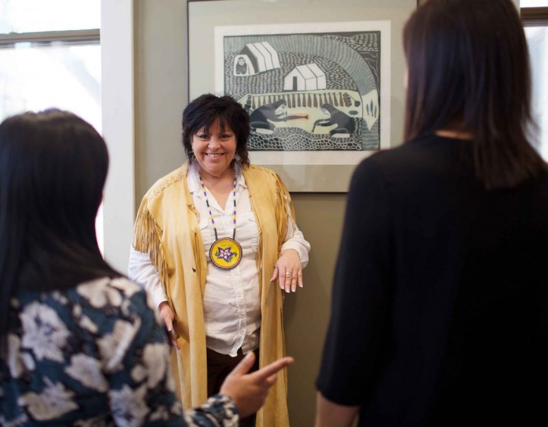 Elder Mary Wilson smiles at the camera while standing beside an Indigenous picture, explaining to two other women, who are facing her.
