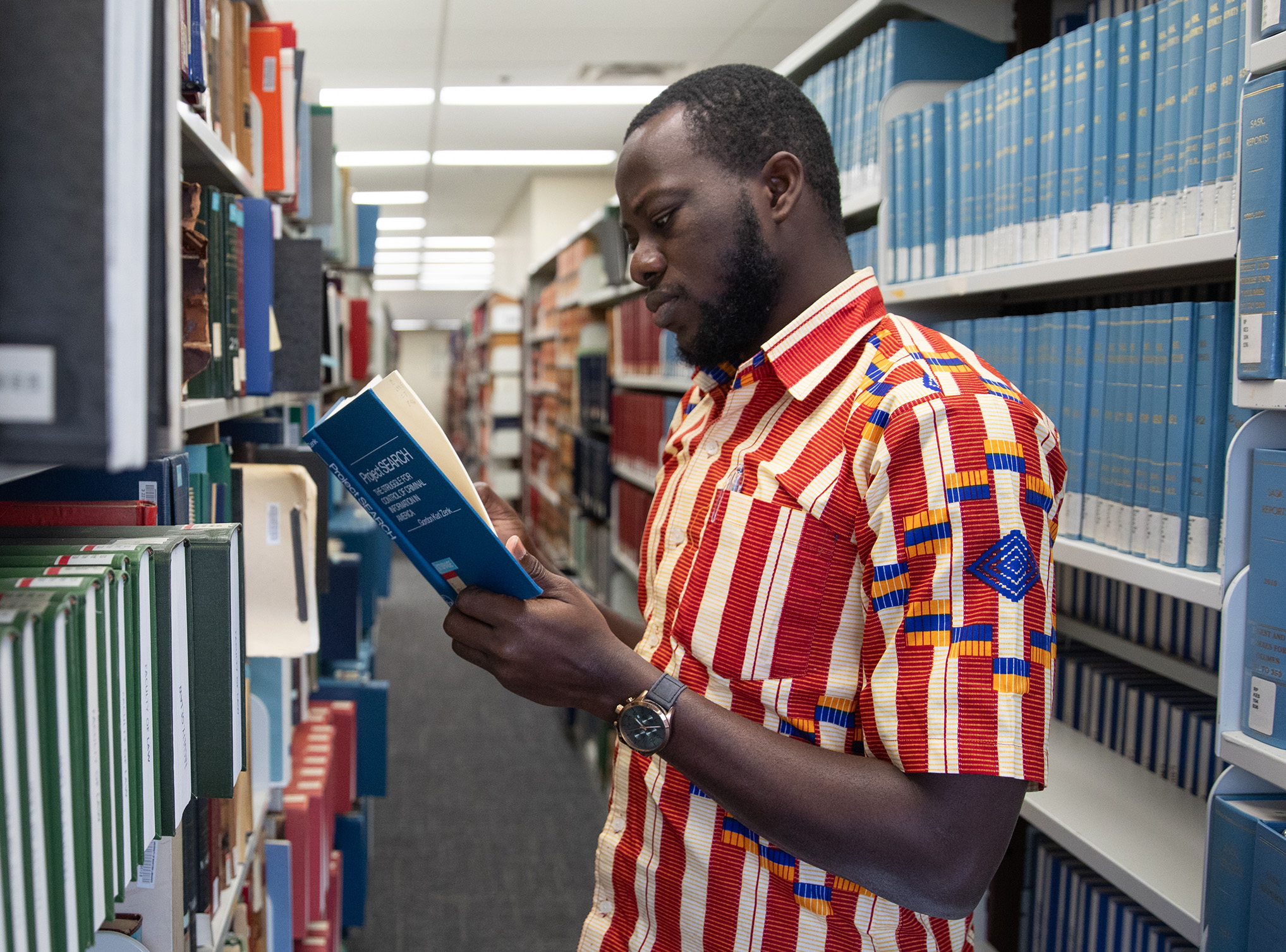 A student in a white and red shirt reads a book in front of library shelves