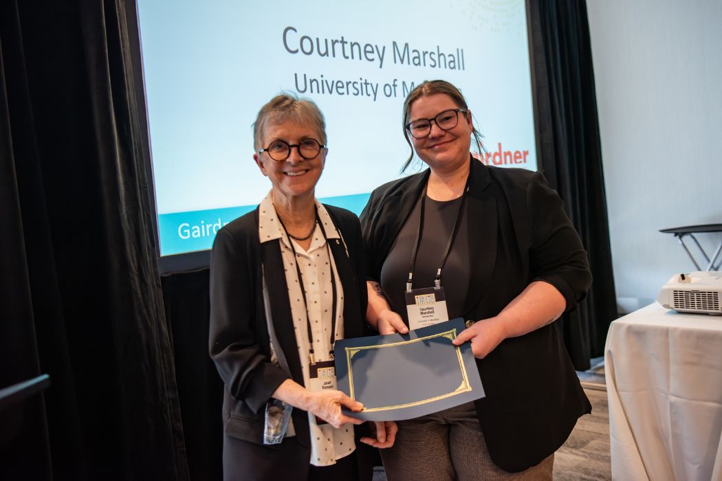 Two people smiling at the camera both holding an award, in the background is text displayed on a screen that says Courtney Marshall, University of Manitoba.