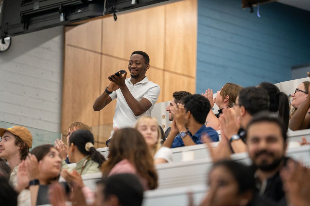 A student standing at an amphitheatre style classroom, surrounded by other students who are smiling and clapping.