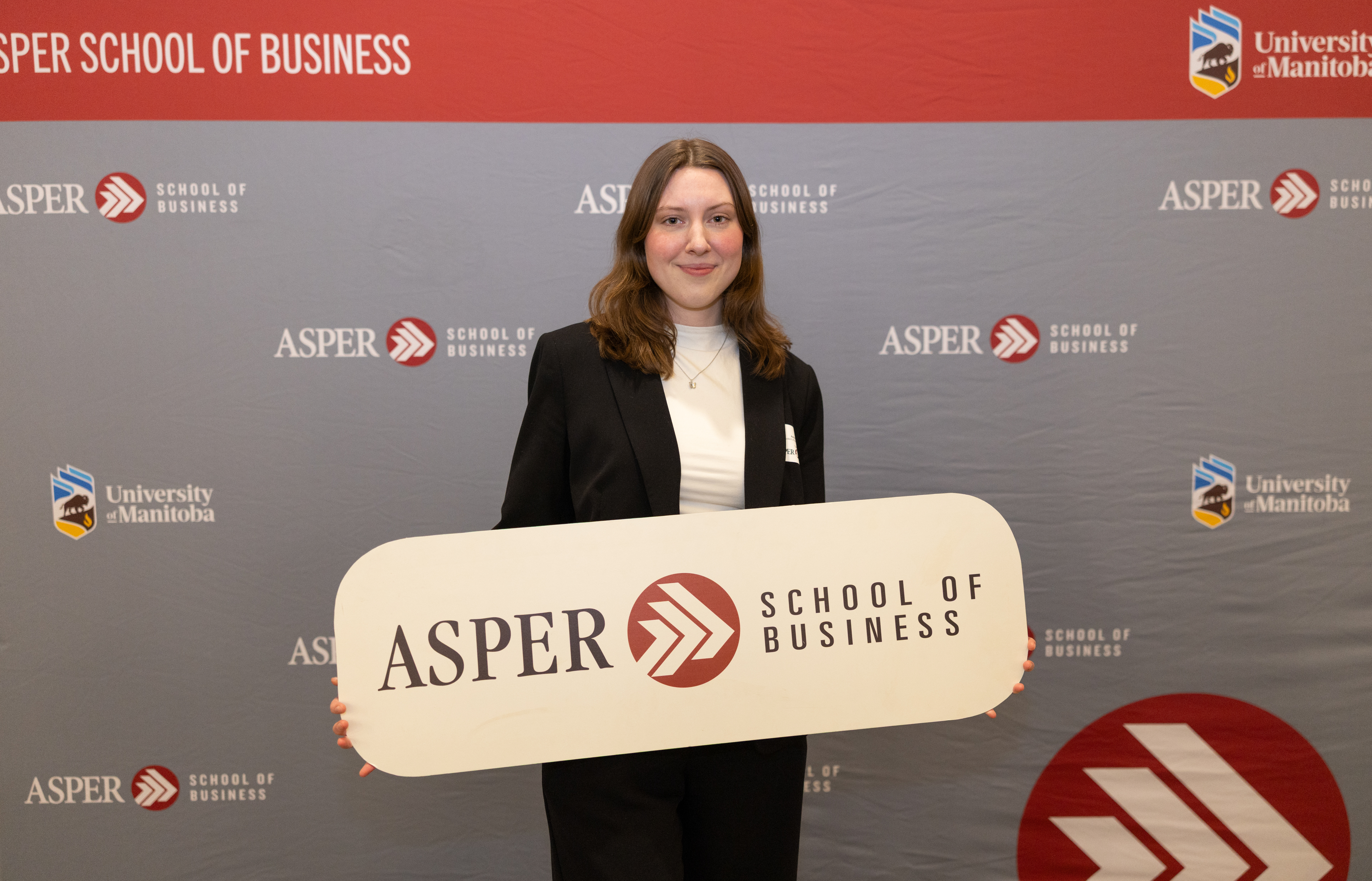 A woman stands in front of an Asper School of Business background holding up a sign, also reading Asper School of Business
