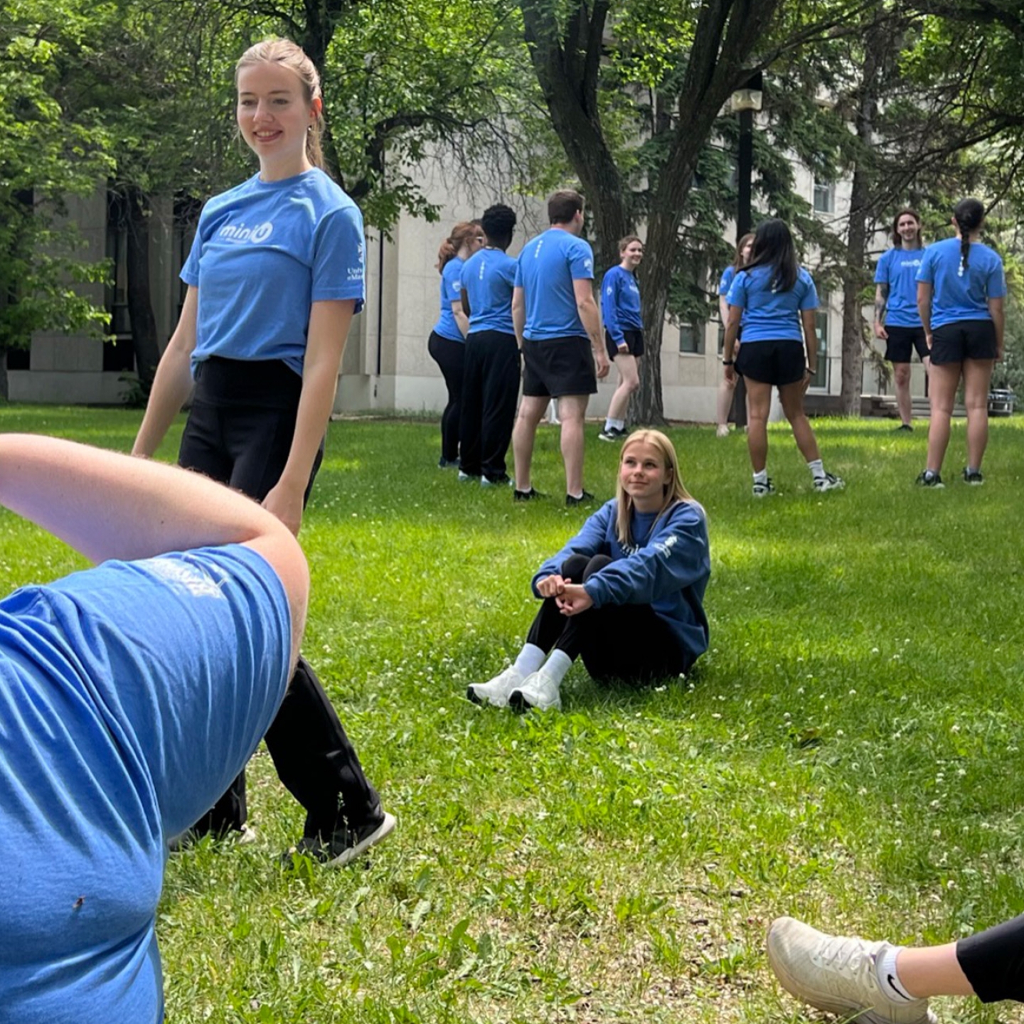 A group of young adults in matching blue shirts stand and sit on a green lawn during Mini U leadersweek in 2025.