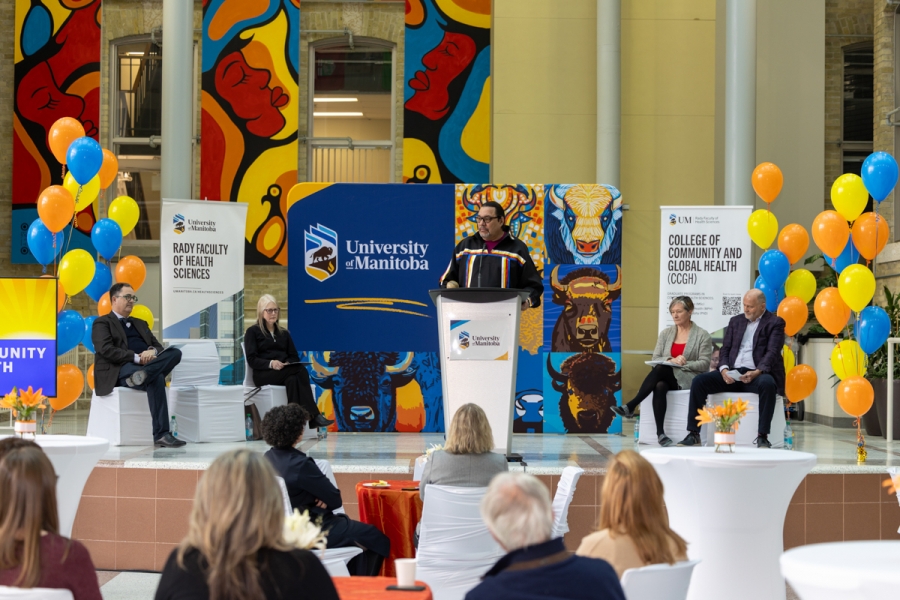 Person speaks onstage while other speakers sit behind. The stage is decorated with balloons and university-branded banner backdrops.