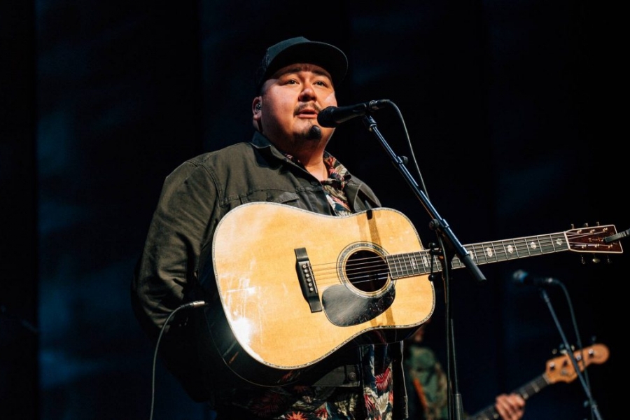 A man in a black baseball cap with a guitar strapped across his chest stands speaking into a microphone