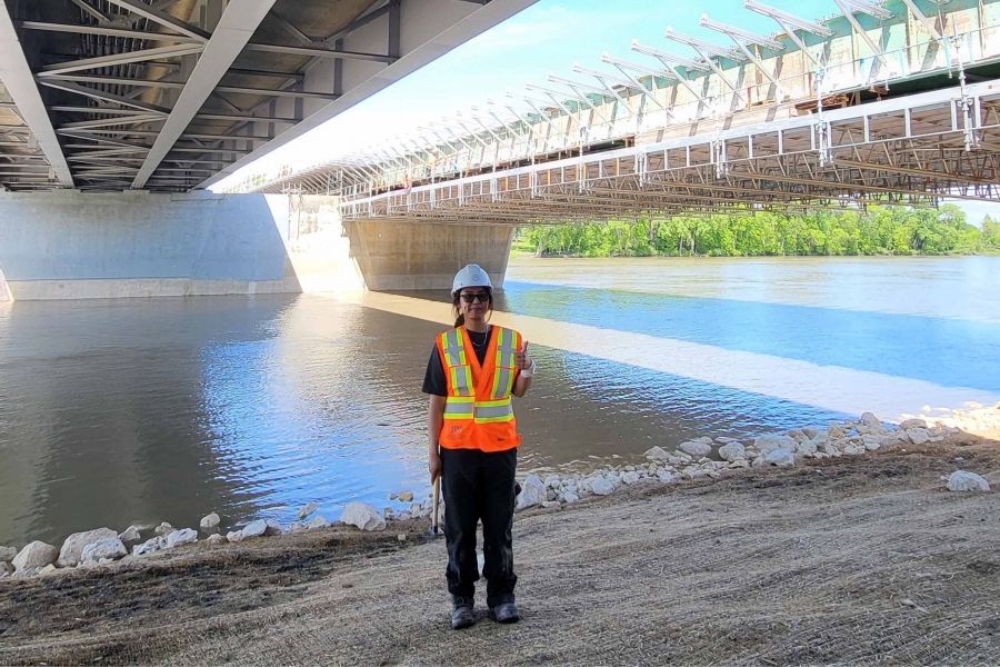 Young woman in construction gear stands under bridge with river in background.