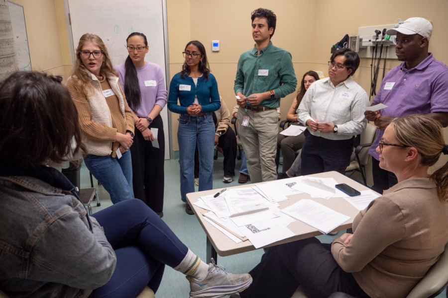 Six students stand while engaging with a Standardized Patient on the left and a facilitator on the right. In the background, other students are seated and observing the interaction.