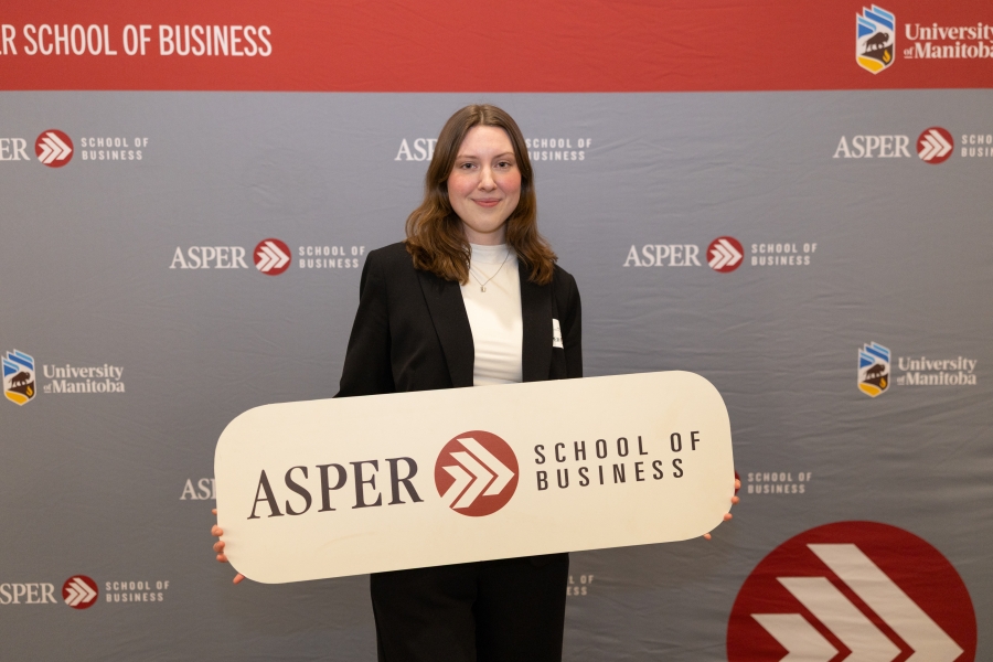 A woman stands in front of an Asper School of Business background holding up a sign, also reading Asper School of Business
