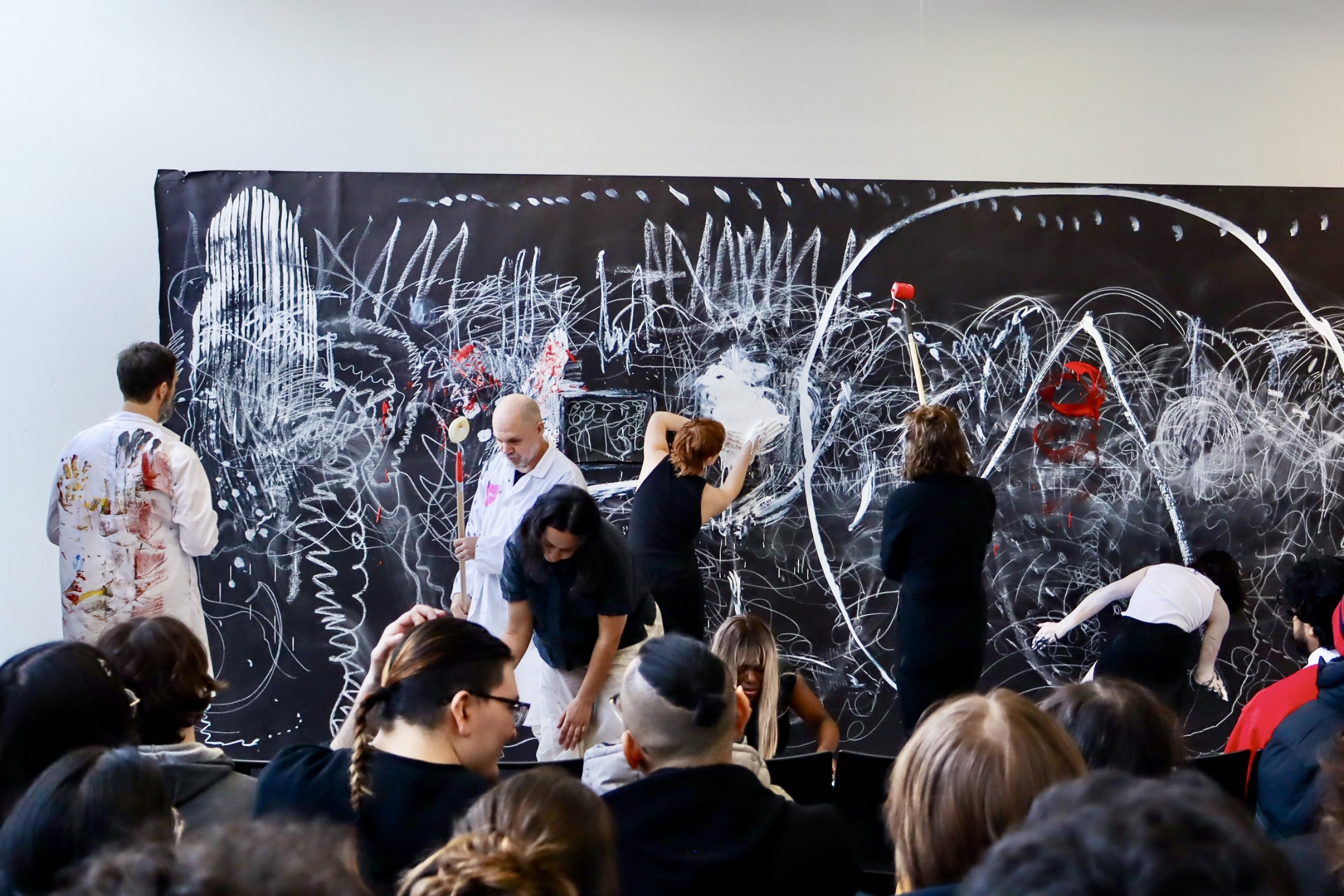 A wide view shows students painting a collaborative artwork during a live performance with an audience present.
