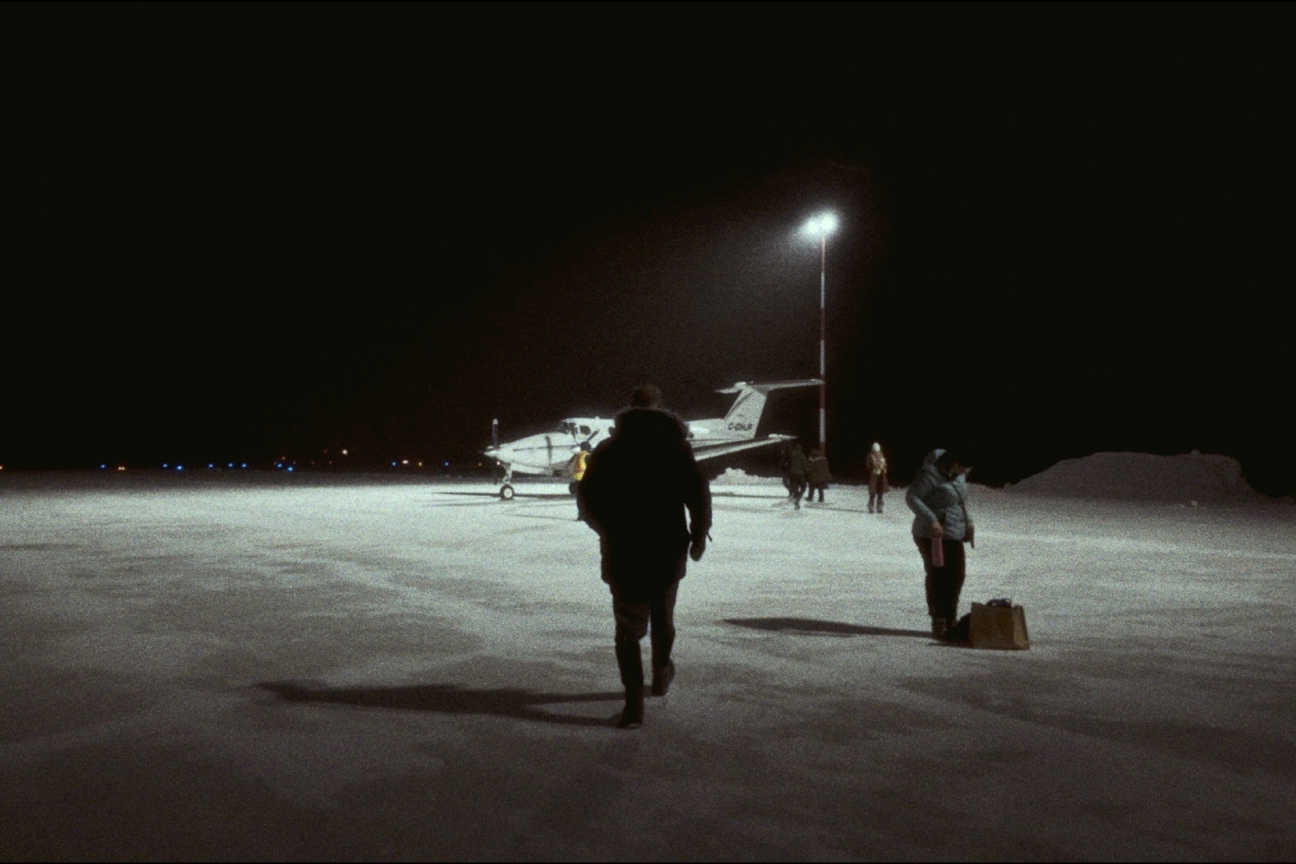 An airplane sits on a snow-covered landing strip lit by one tall spotlight. A person walks towards it. about 4 other people stand nearby, waiting to board.