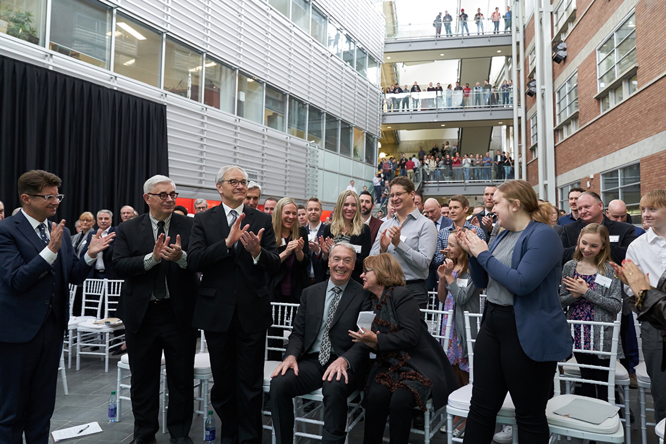 A crowded atrium gives a standing ovation to Gerry Price while he is seated.