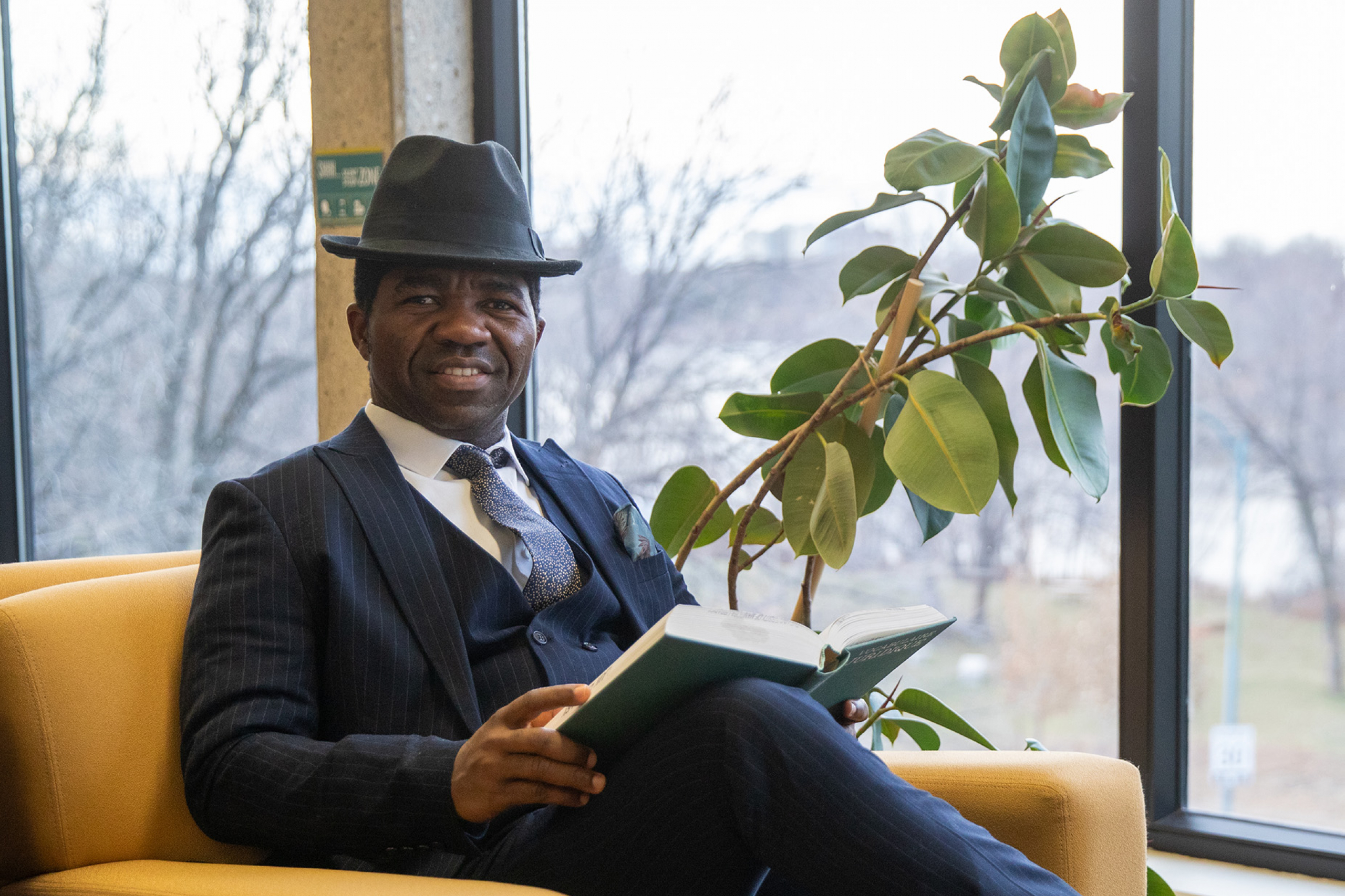 A man in a suit wears a hat and smiles at the camera holding a book, sitting in a chair in front of a large window.