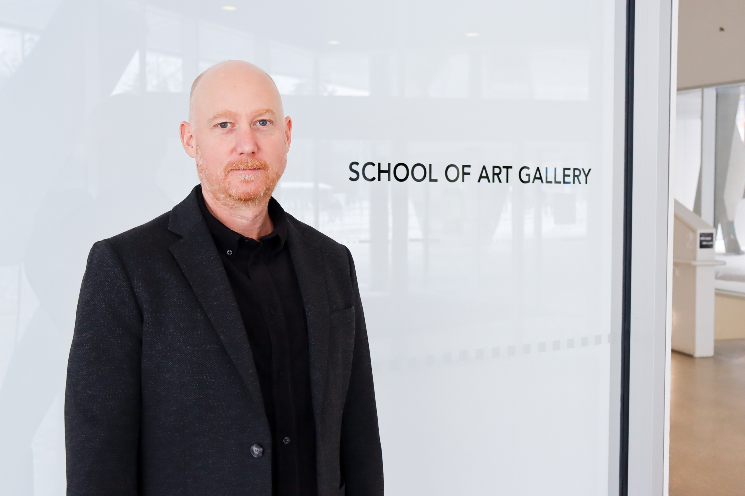 Derek Dunlop stands in a bright hallway beside a glass wall reading “School of Art Gallery,” wearing a dark blazer and black shirt.