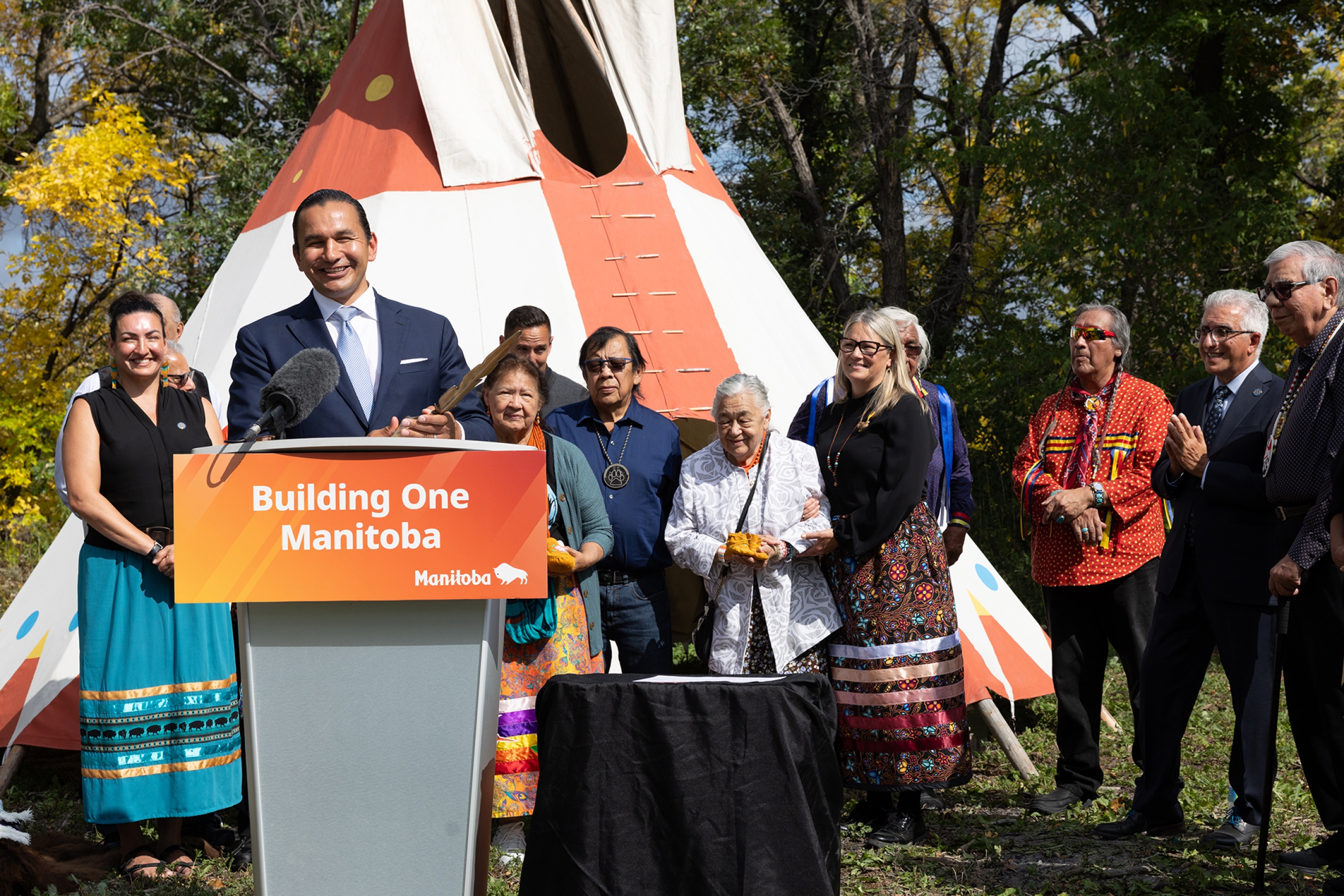 Premier Wab Kinew standing at a podium with Elders, Survivors and UM staff standing behind him