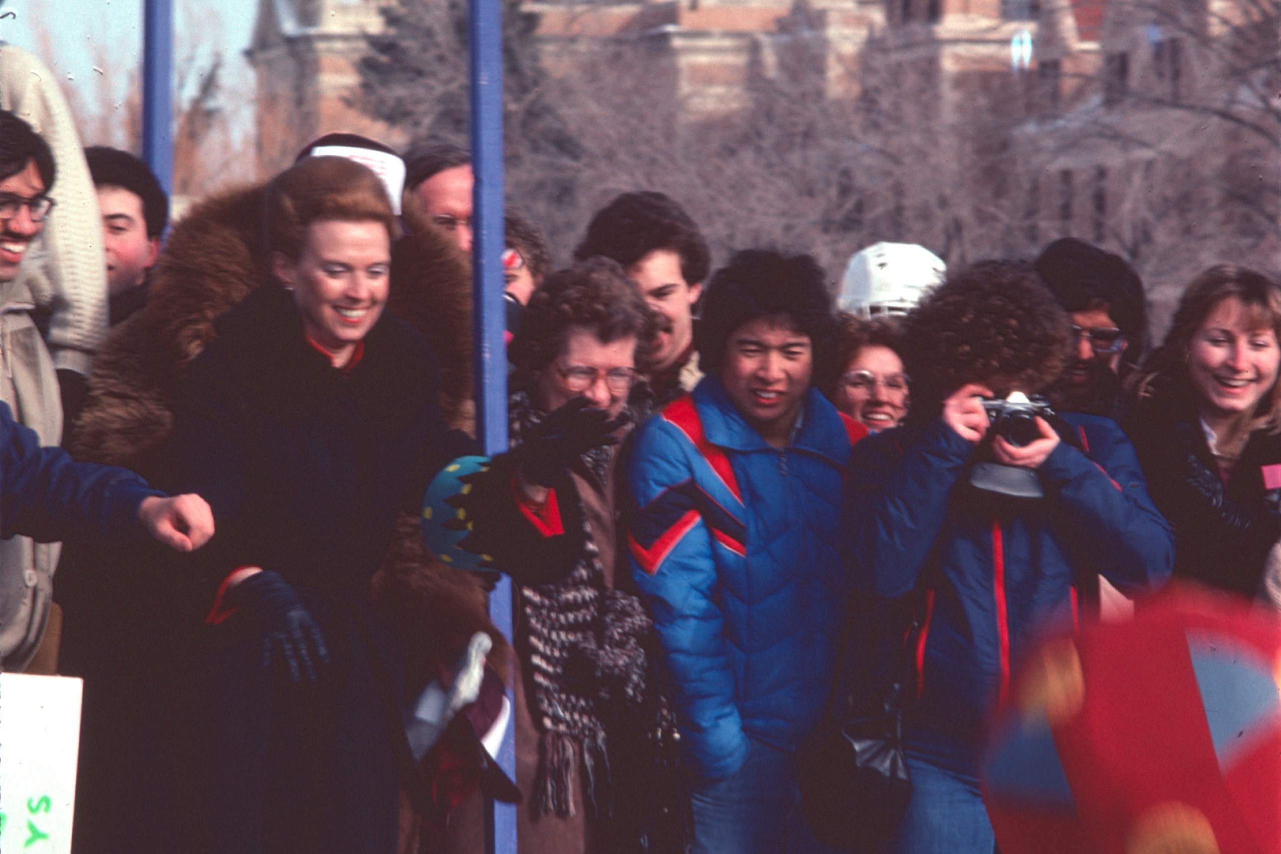 A group of people in winter clothing stand outdoors near a blue metal frame, with snowy ground and an old building in the background. 