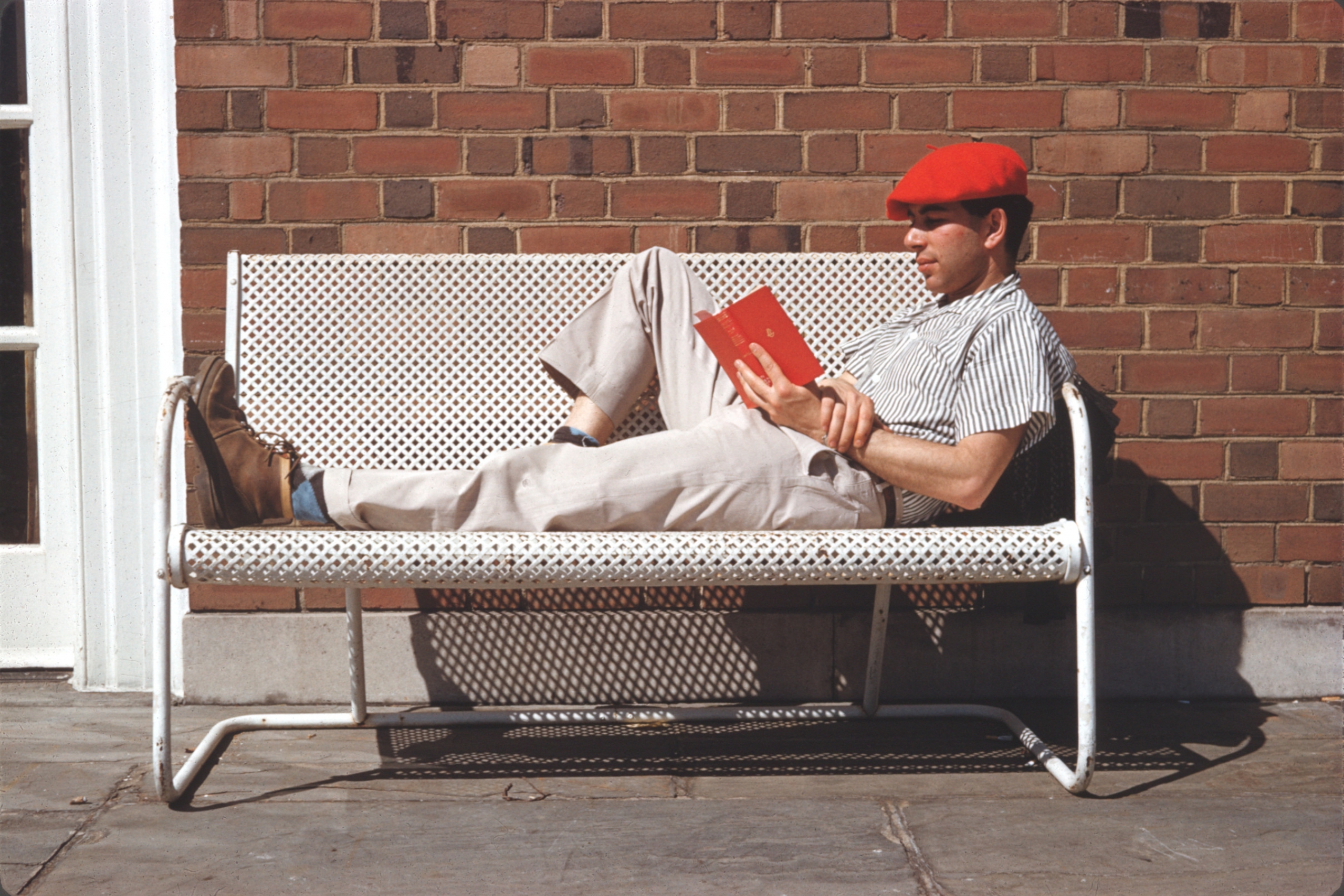 A man in a red cap lies on a white perforated bench against a brick wall, reading a red book.