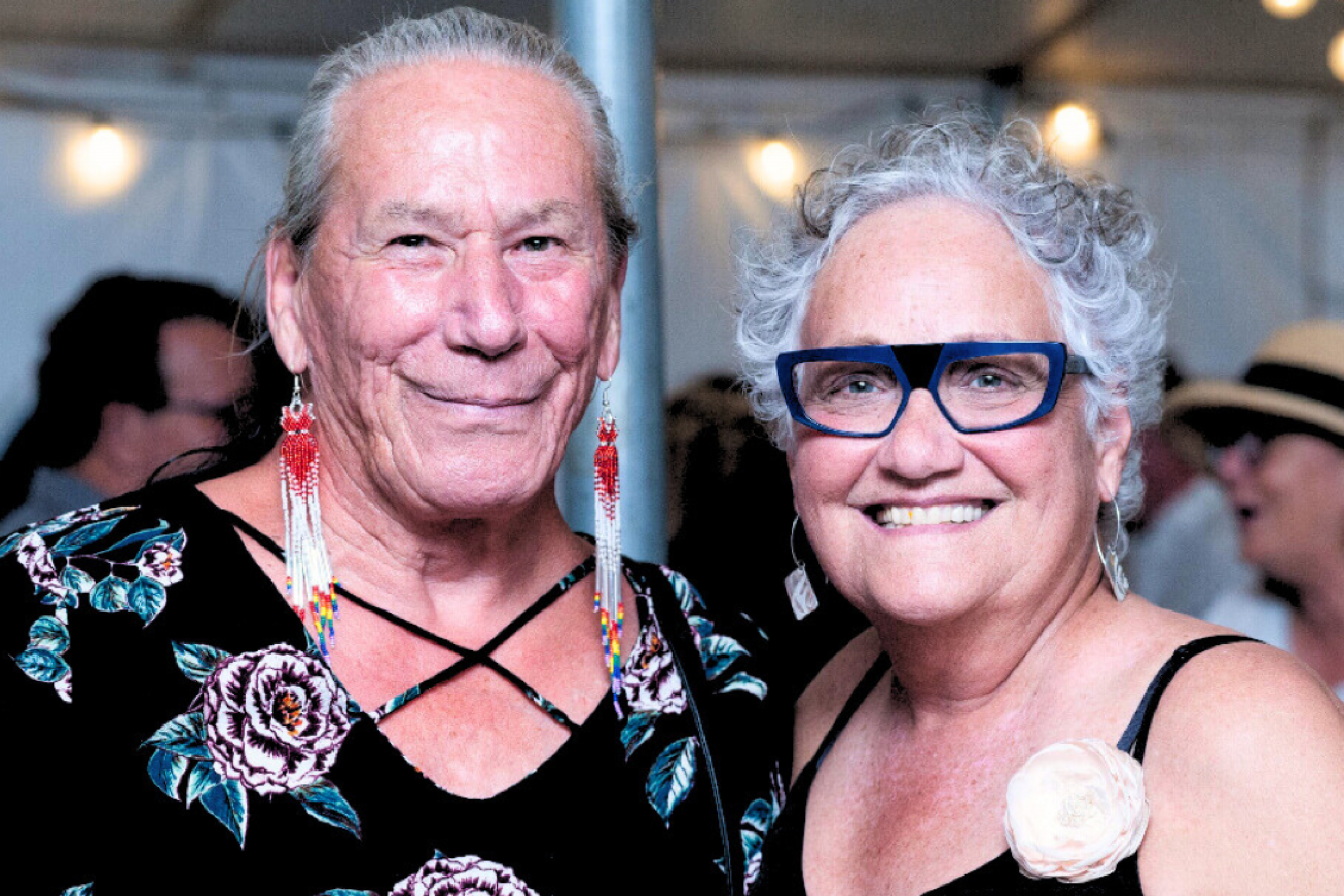 Two Two-Spirit people smiling together at a community event under string lights.