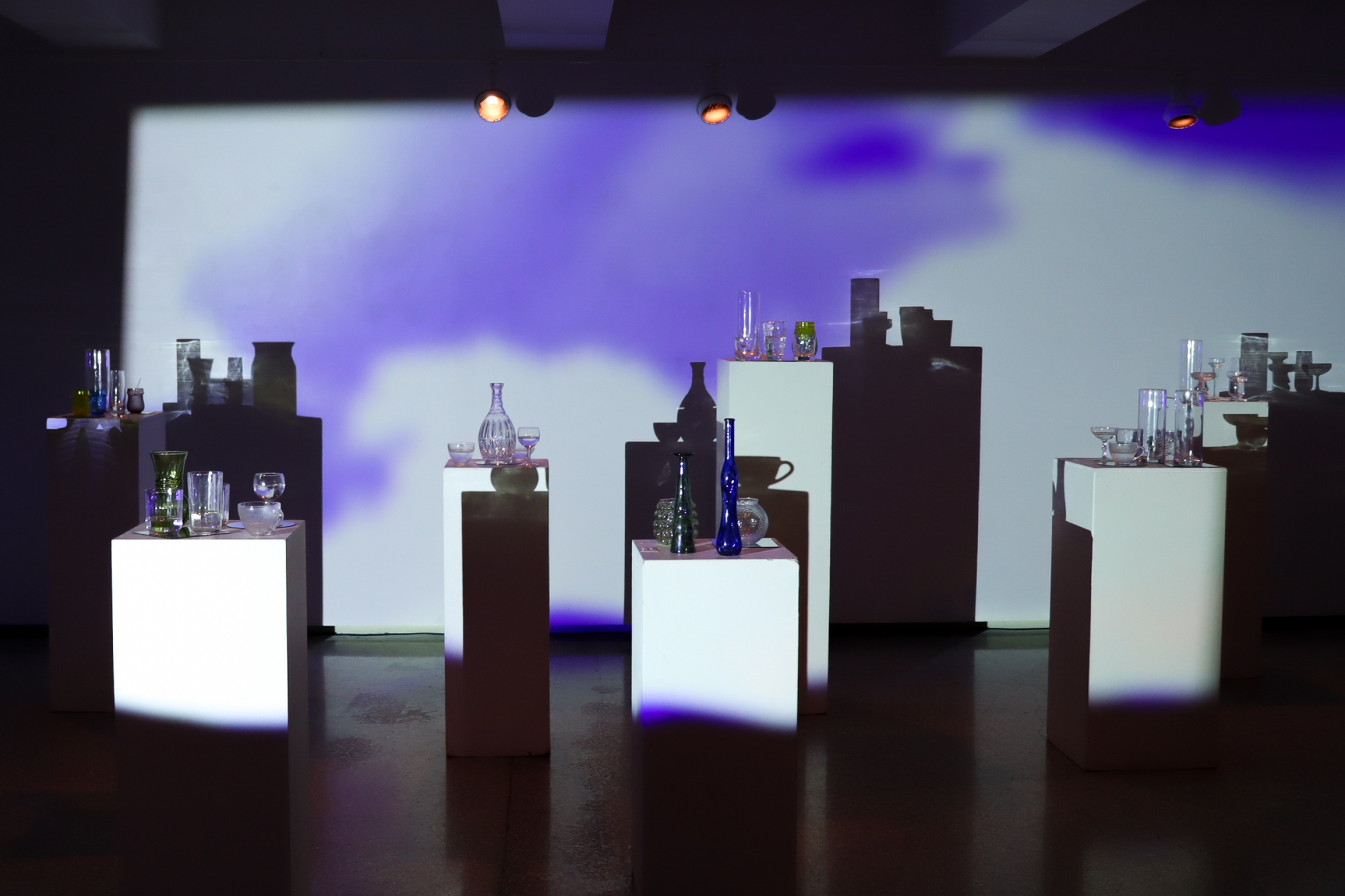 Wide view of ASTRAL installation with glass vessels on white plinths, casting dramatic shadows under shifting blue-violet projection.