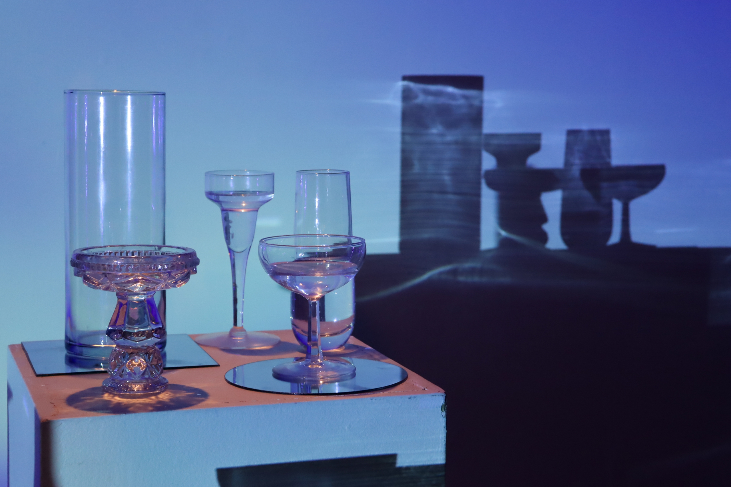 Close-up of glass vessels on a plinth lit in cool blue, with their tall shadows layered against projected light on the wall behind.