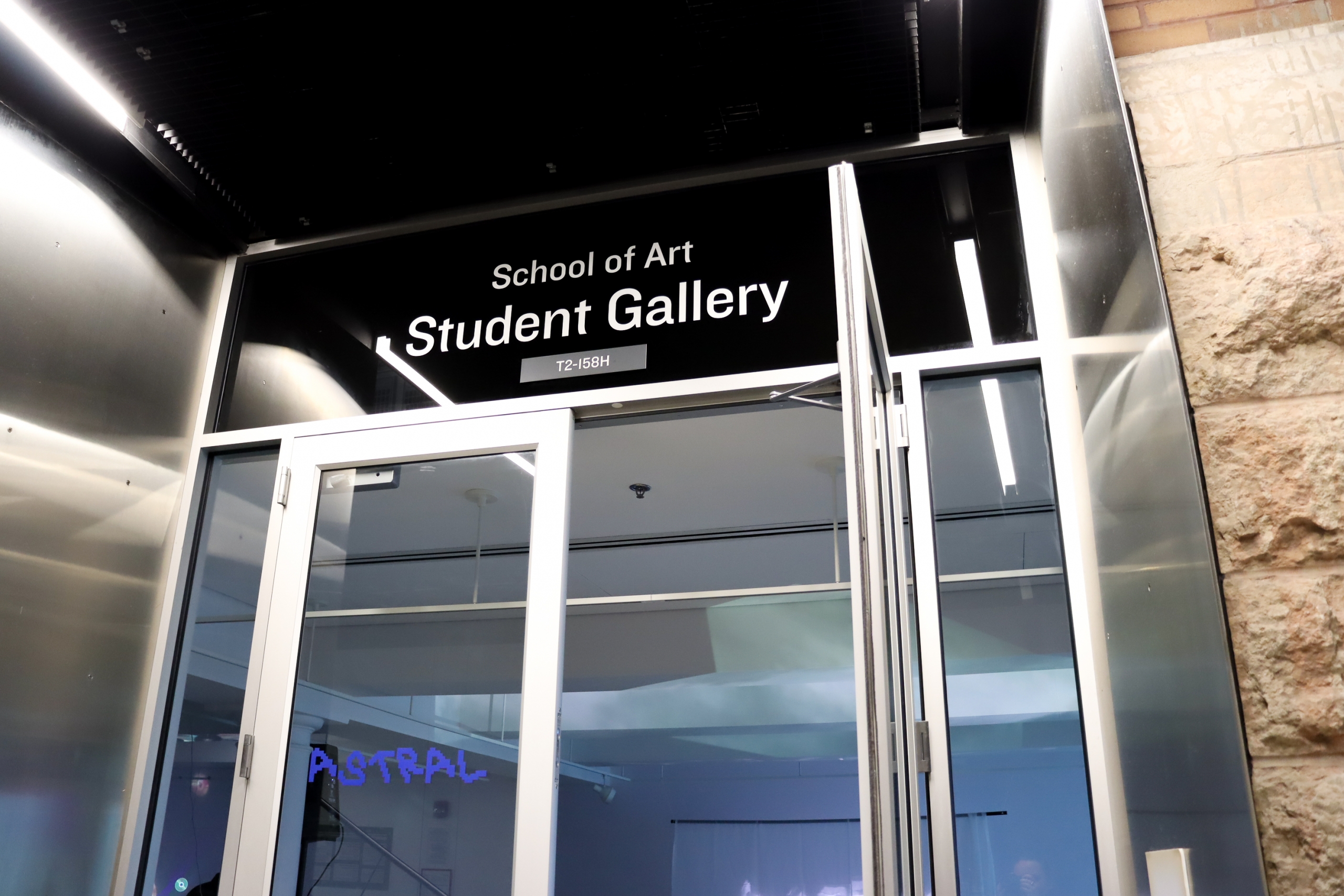 Exterior view of the School of Art Student Gallery entrance with the doors open and “ASTRAL” glowing in blue inside.