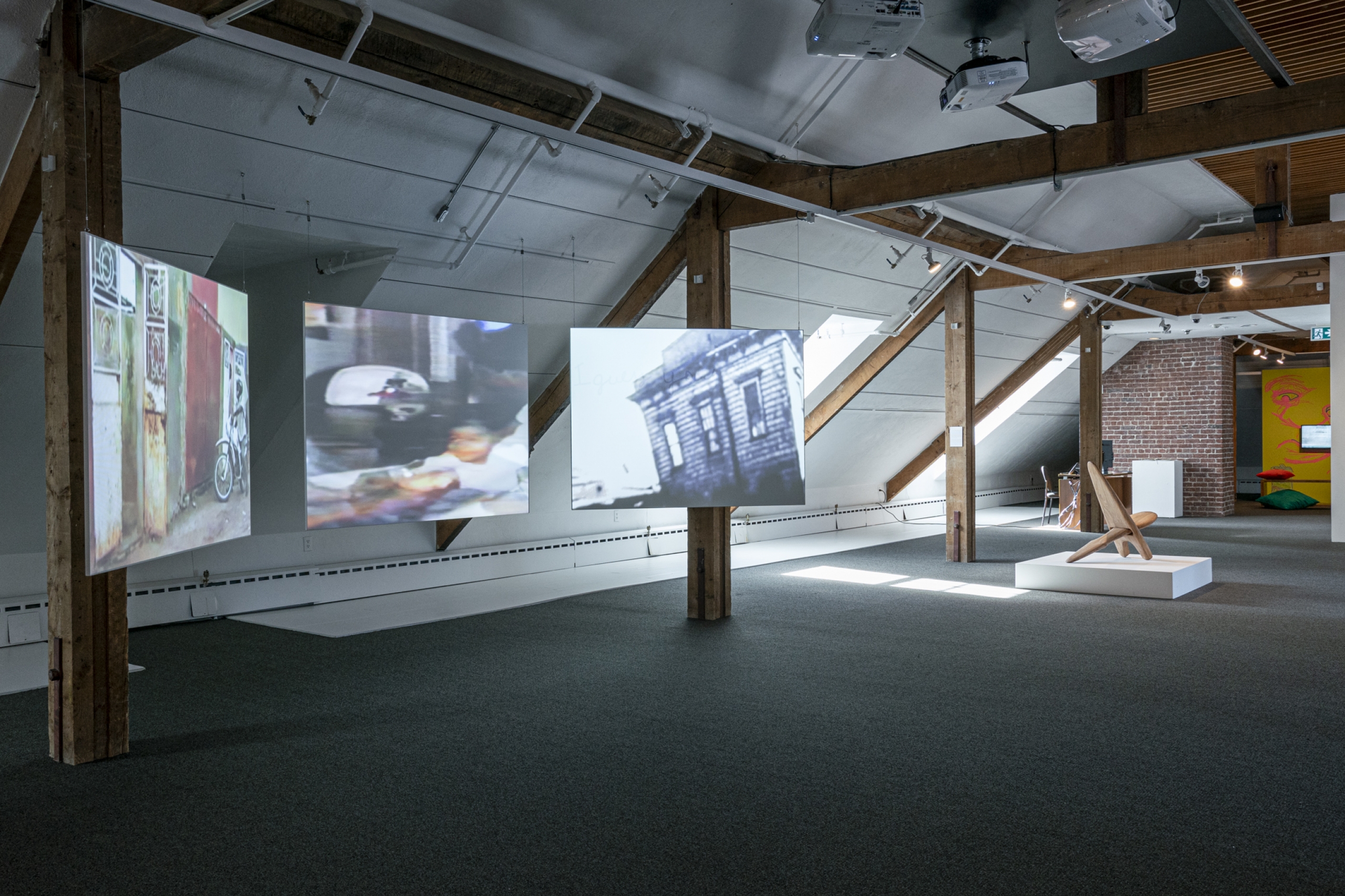 An art exhibit featuring three suspended screens showing blurred video projections and a wooden sculptural chair on a low pedestal.