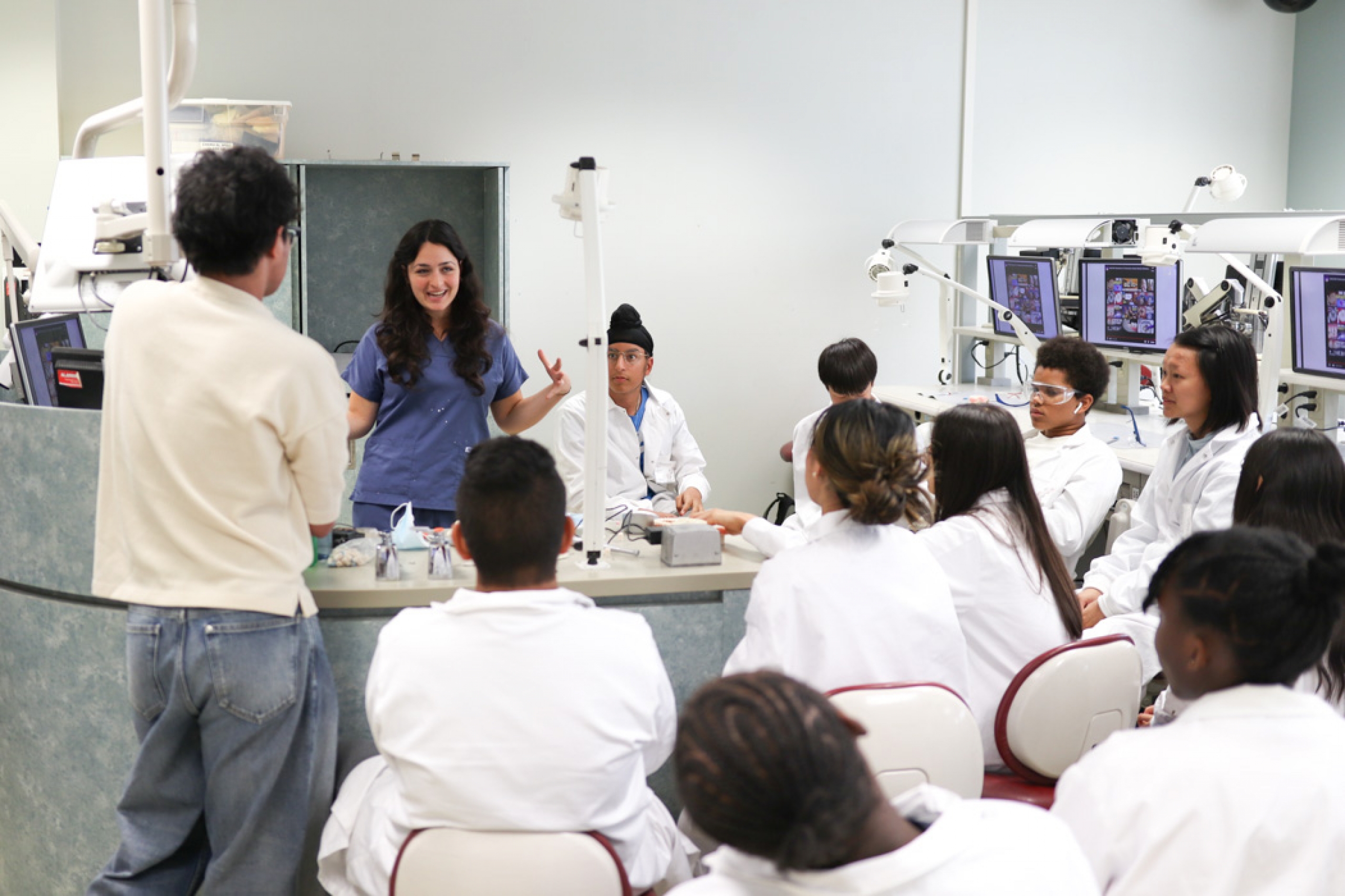 A group of campers wearing lab coats sit in the dentistry lab listening to one of the dentistry workshop instructors.
