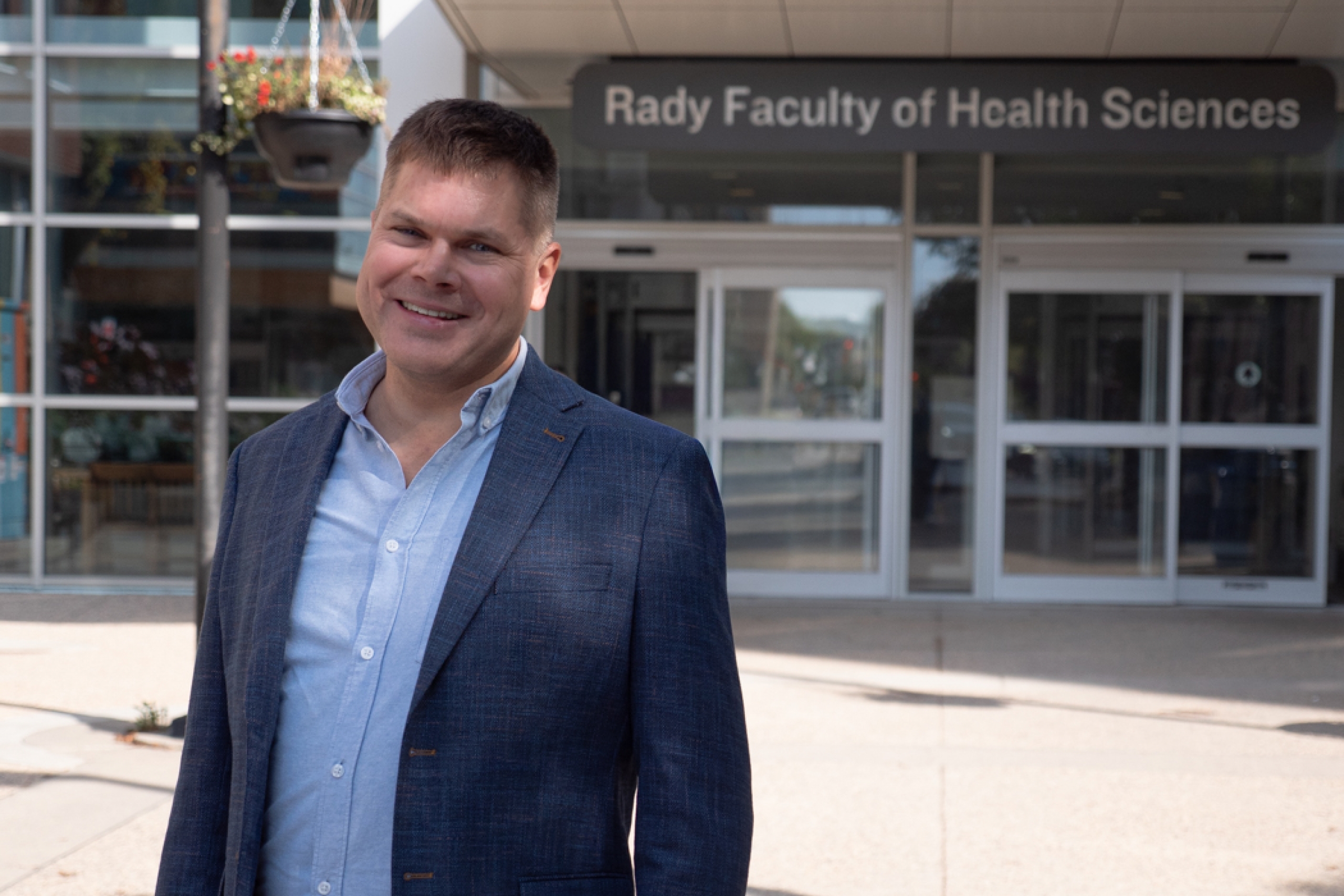 Barret Monchka smiles at the camera while standing outside the Brodie Centre. In the background, the entrance to the Rady Faculty of Health Sciences is visible.