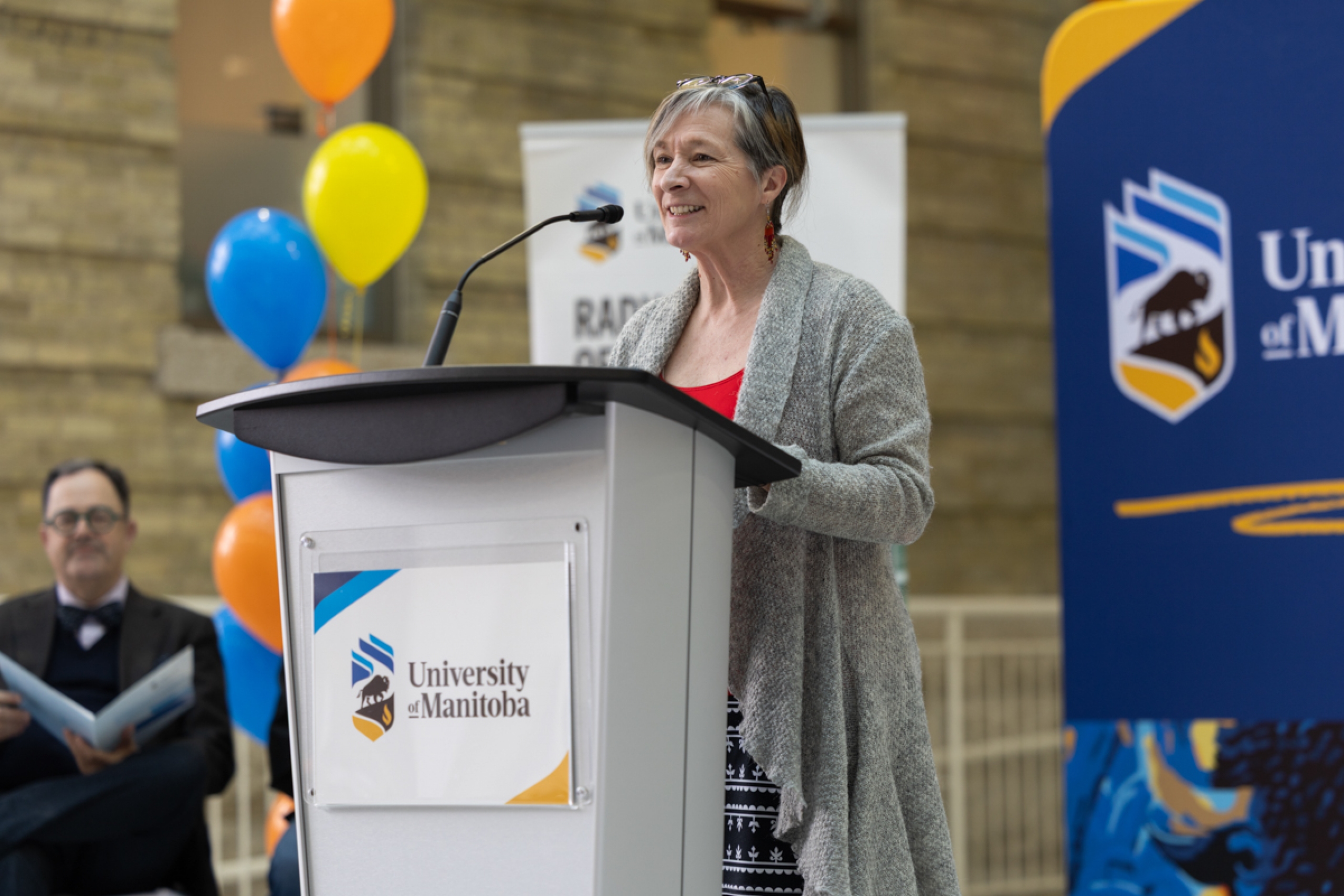 Dr. Josée Lavoie speaks at a podium onstage. The stage is decorated with balloons and university-branded banner backdrops.