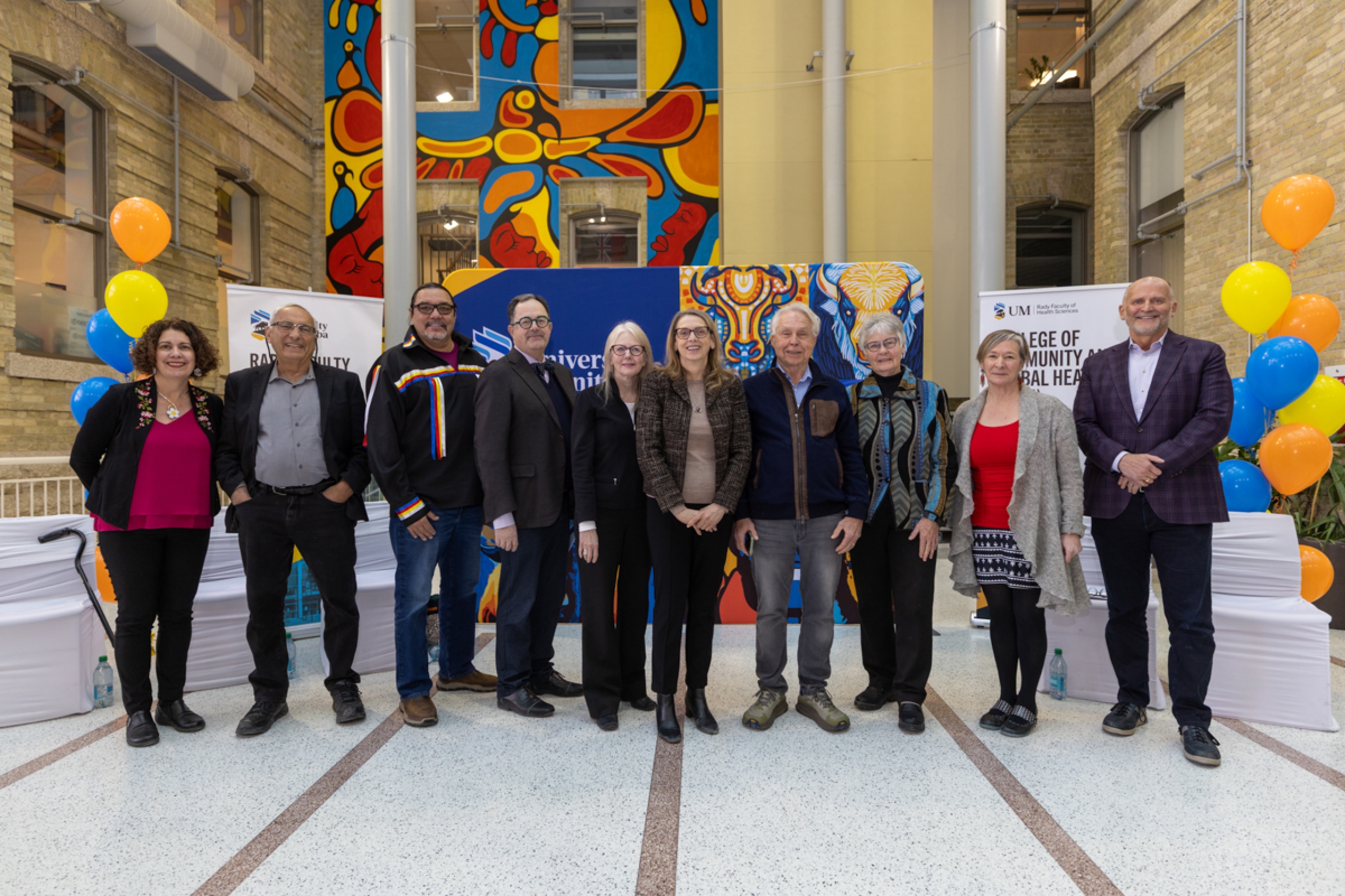 A group of ten people onstage smile at the camera. The stage is decorated with balloons and university-branded banner backdrops.
