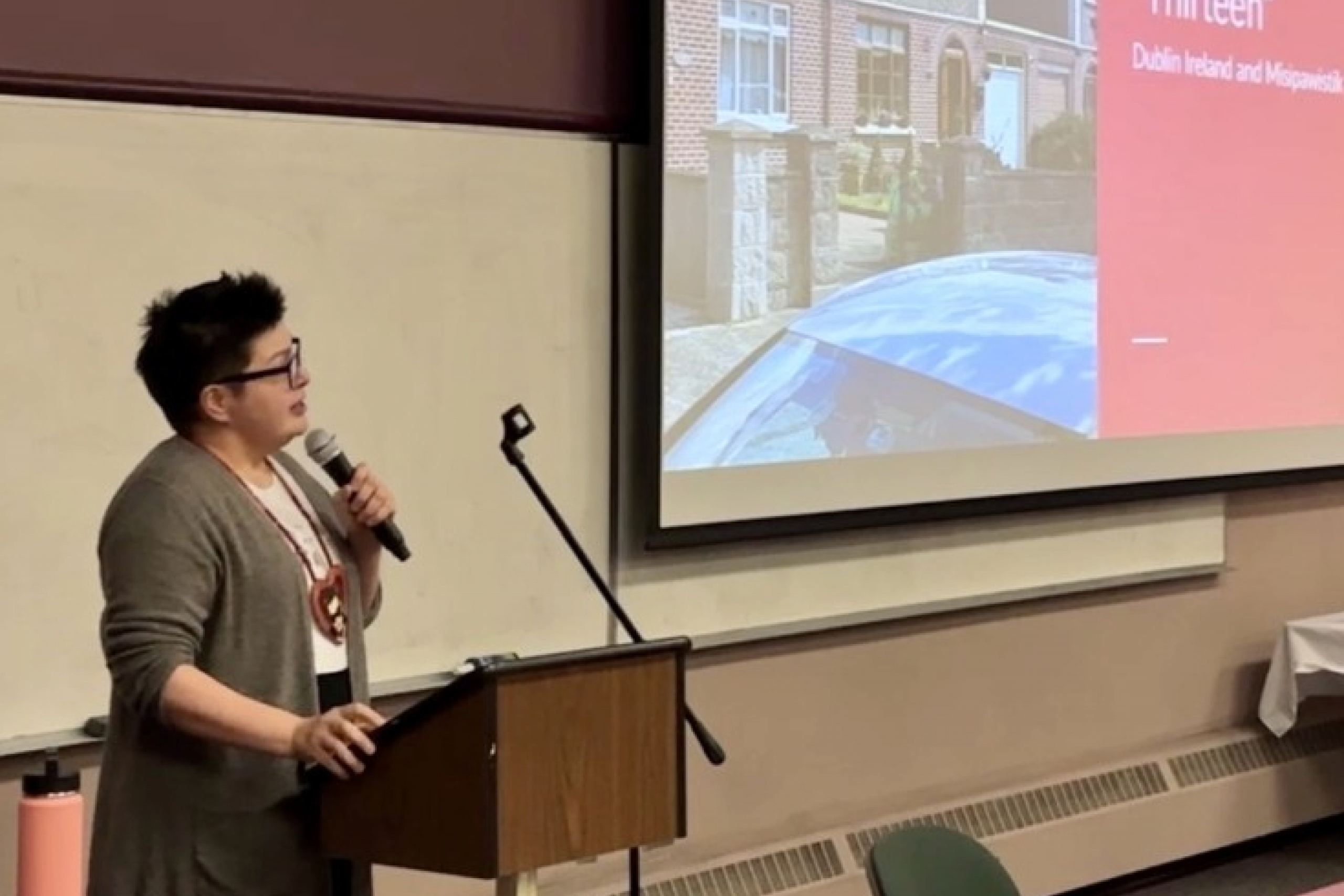 Woman presenting at the front of a classroom. She is looking up at a large screen.