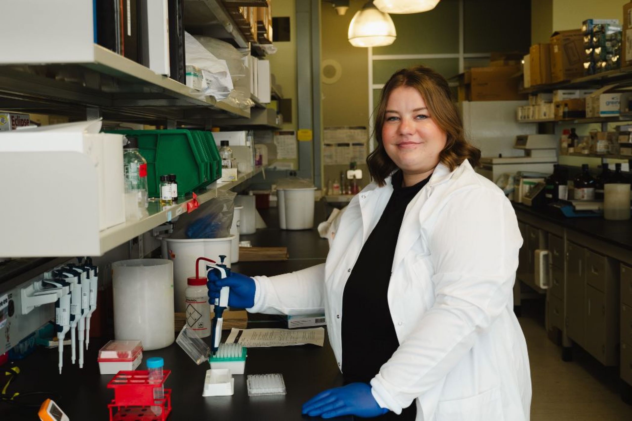 UM researcher, Courtney Marshall in a lab wearing a white lab coat and holding a lab apparatus while smiling at the camera.