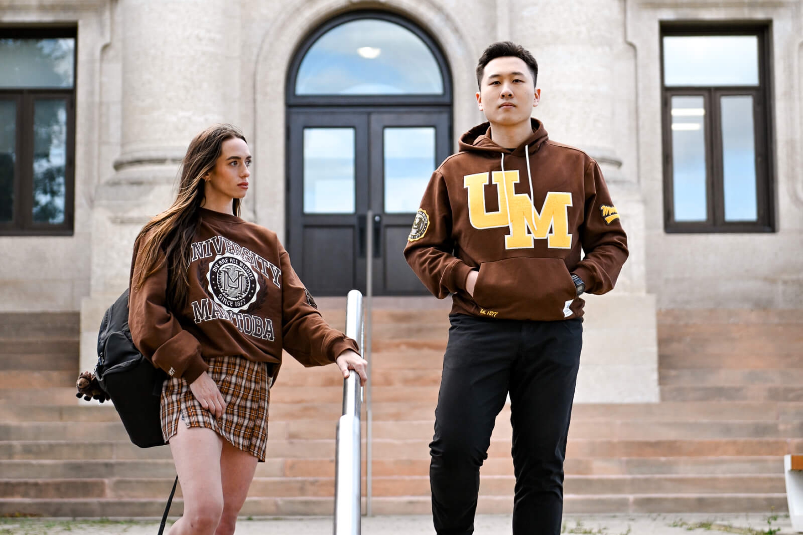 Two students stand on the steps of the Admin building wearing new Mondetta and UM clothing