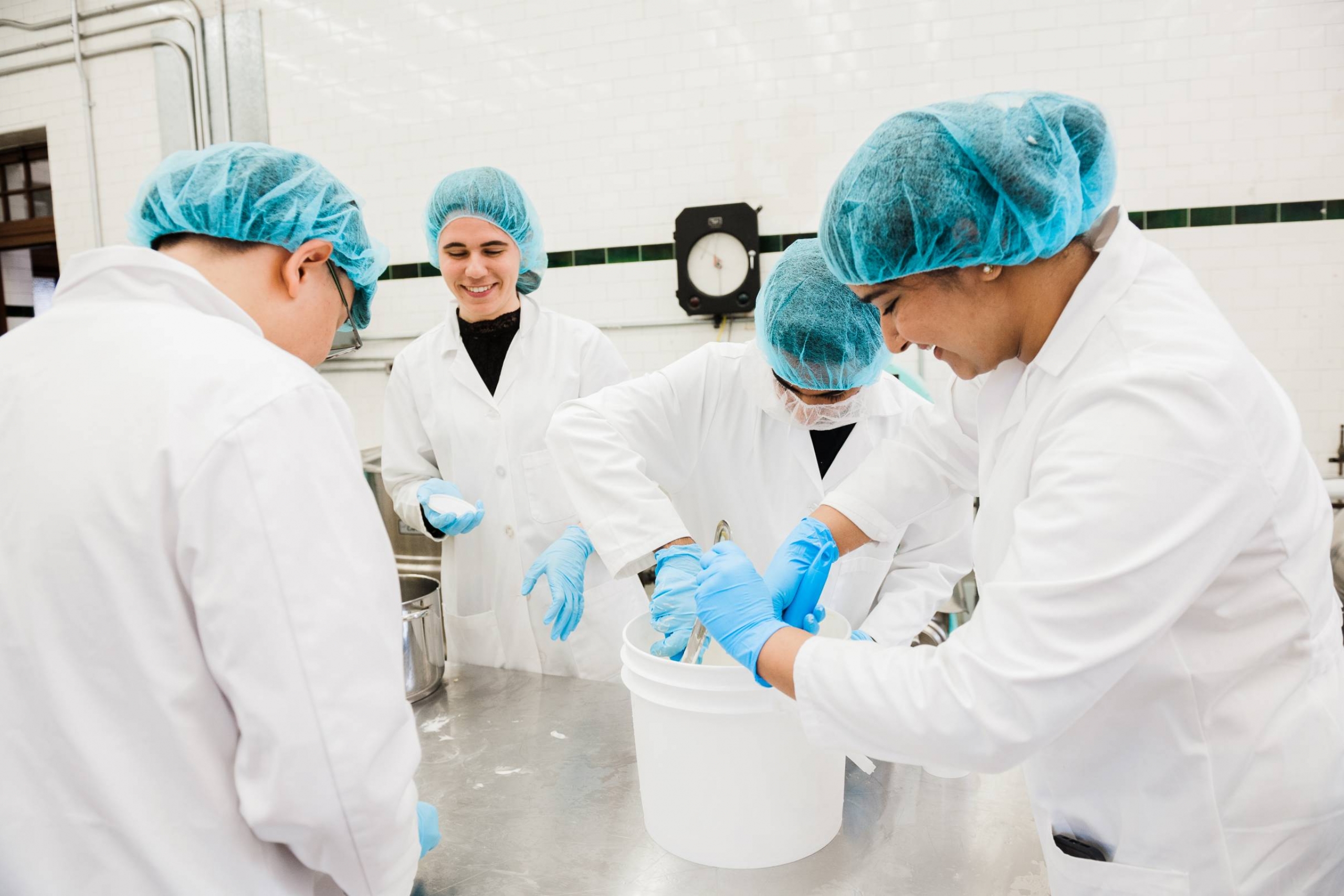 Students in UM's dairy plant are stood around a table, each making ice cream, wearing white lab coats and teal hair nets