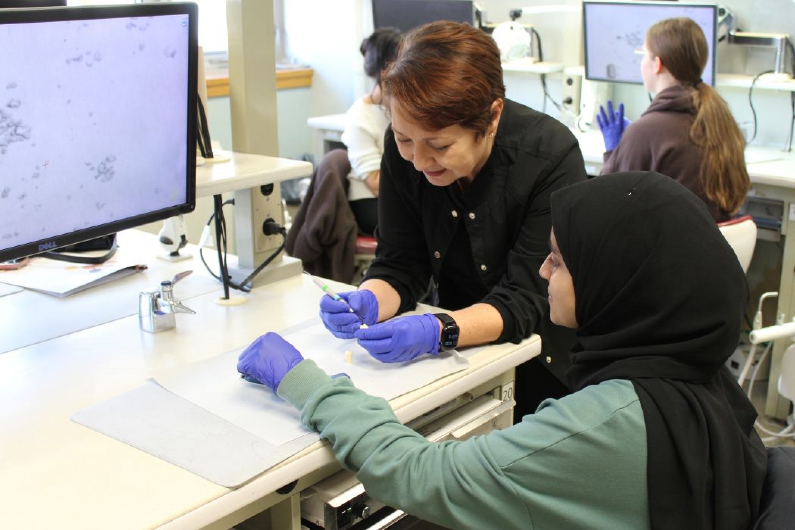 An instructor demonstrates a dental technique with a model tooth and a dental instrument as a student watches.