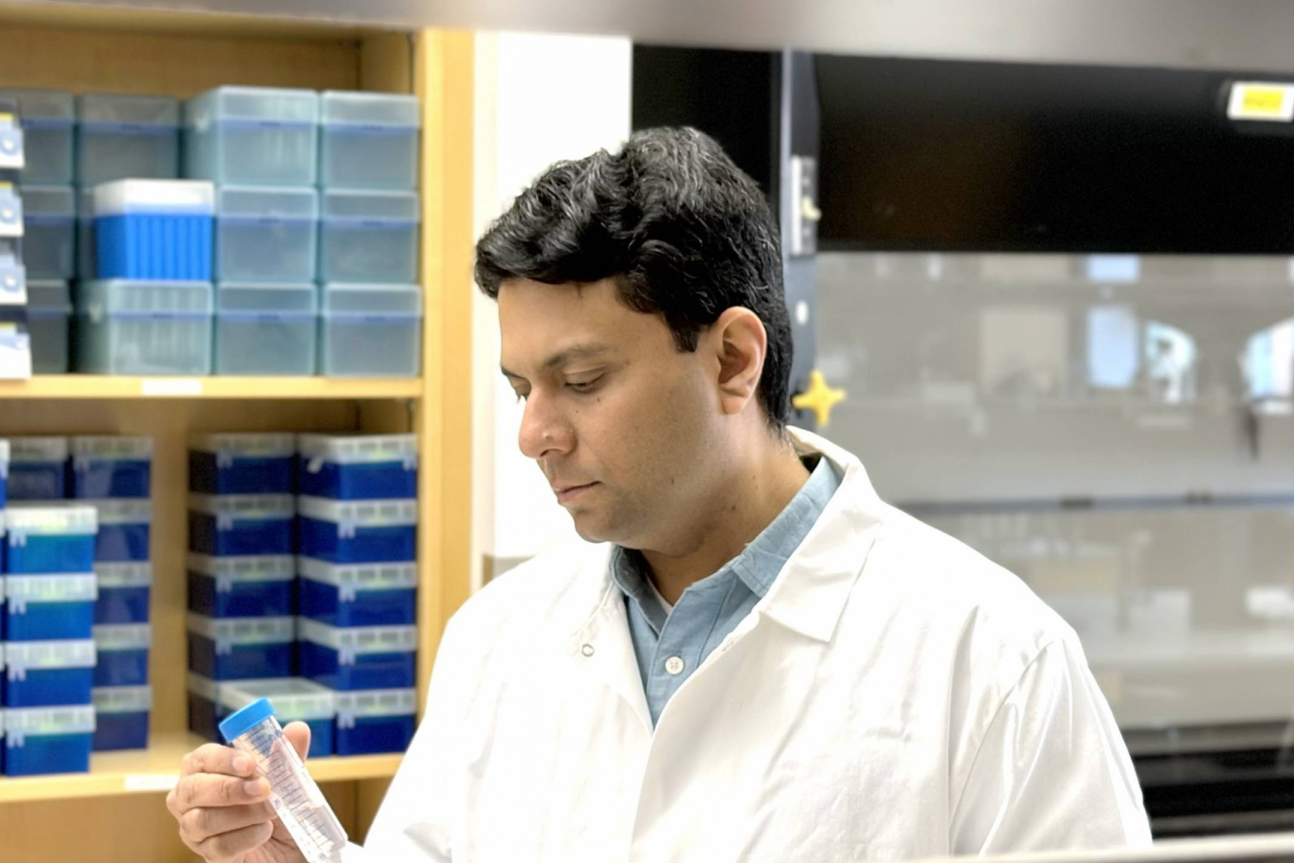Dr. Abdullah Maruf, wearing a white lab coat, in a laboratory holding a test tube. Shelves with scientific equipment are visible in the background.