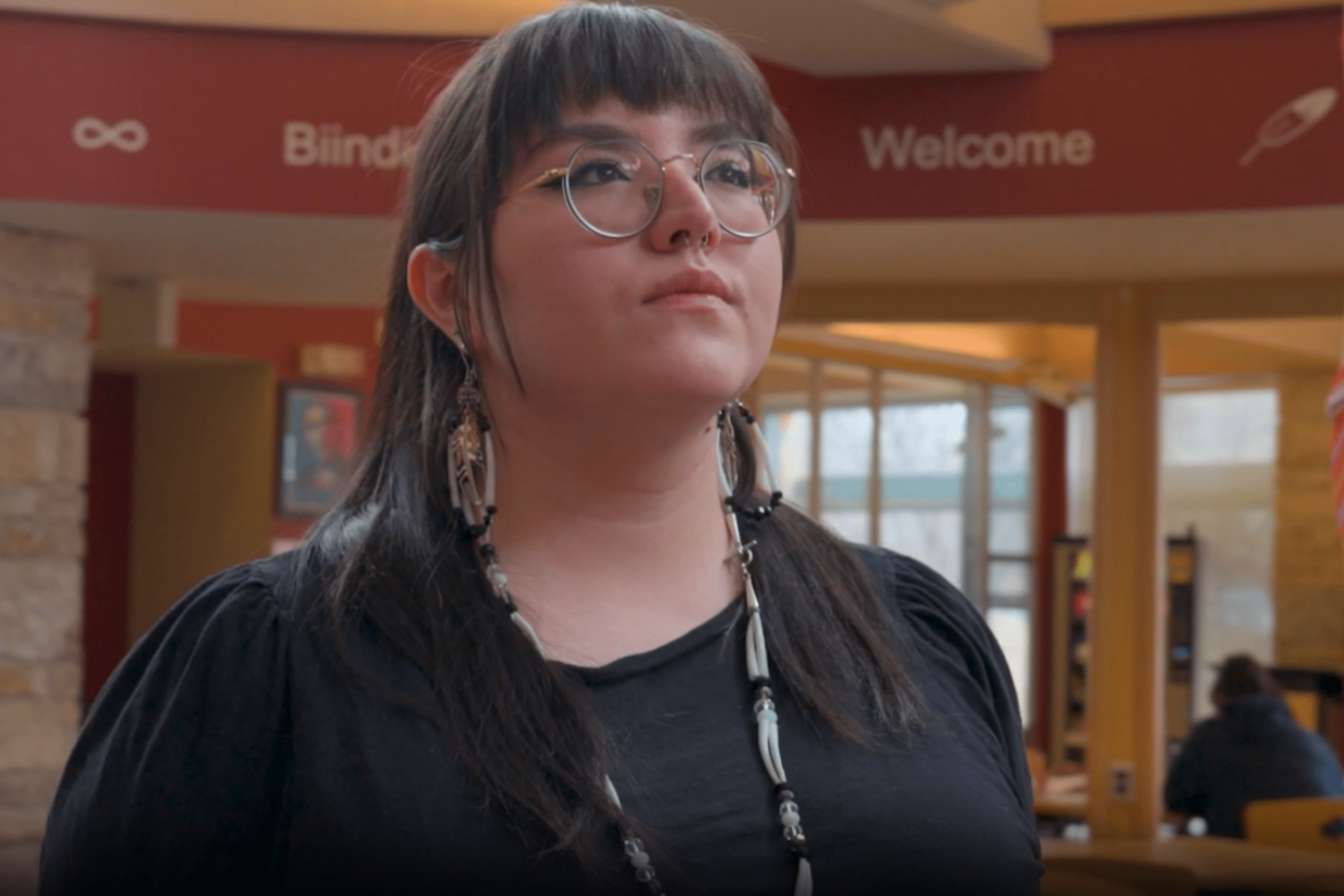 Student stands in Migizii Agamik - Bald Eagle lodge beside a pole wrapped in a Métis sash.