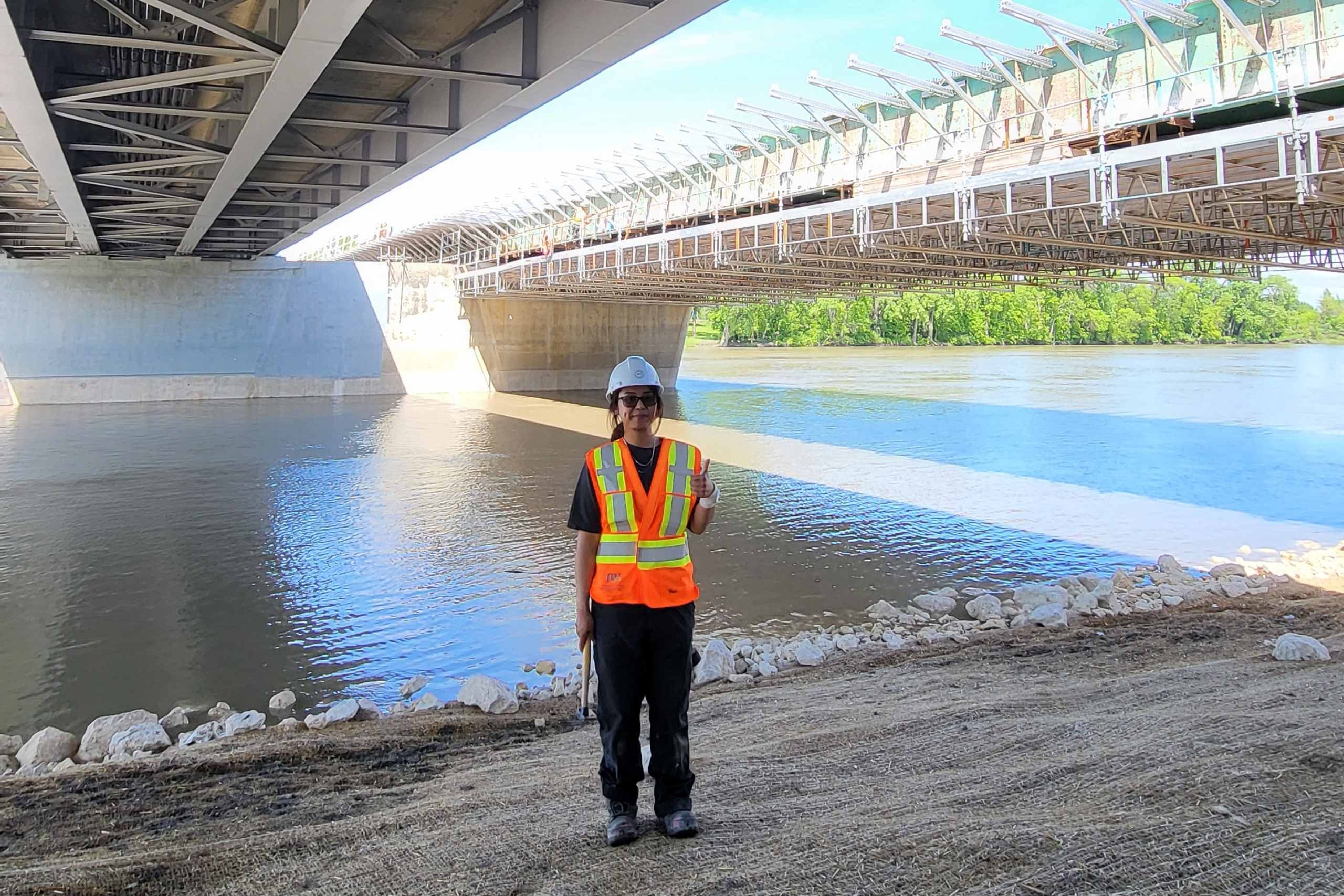 Young woman in construction gear stands under bridge with river in background.