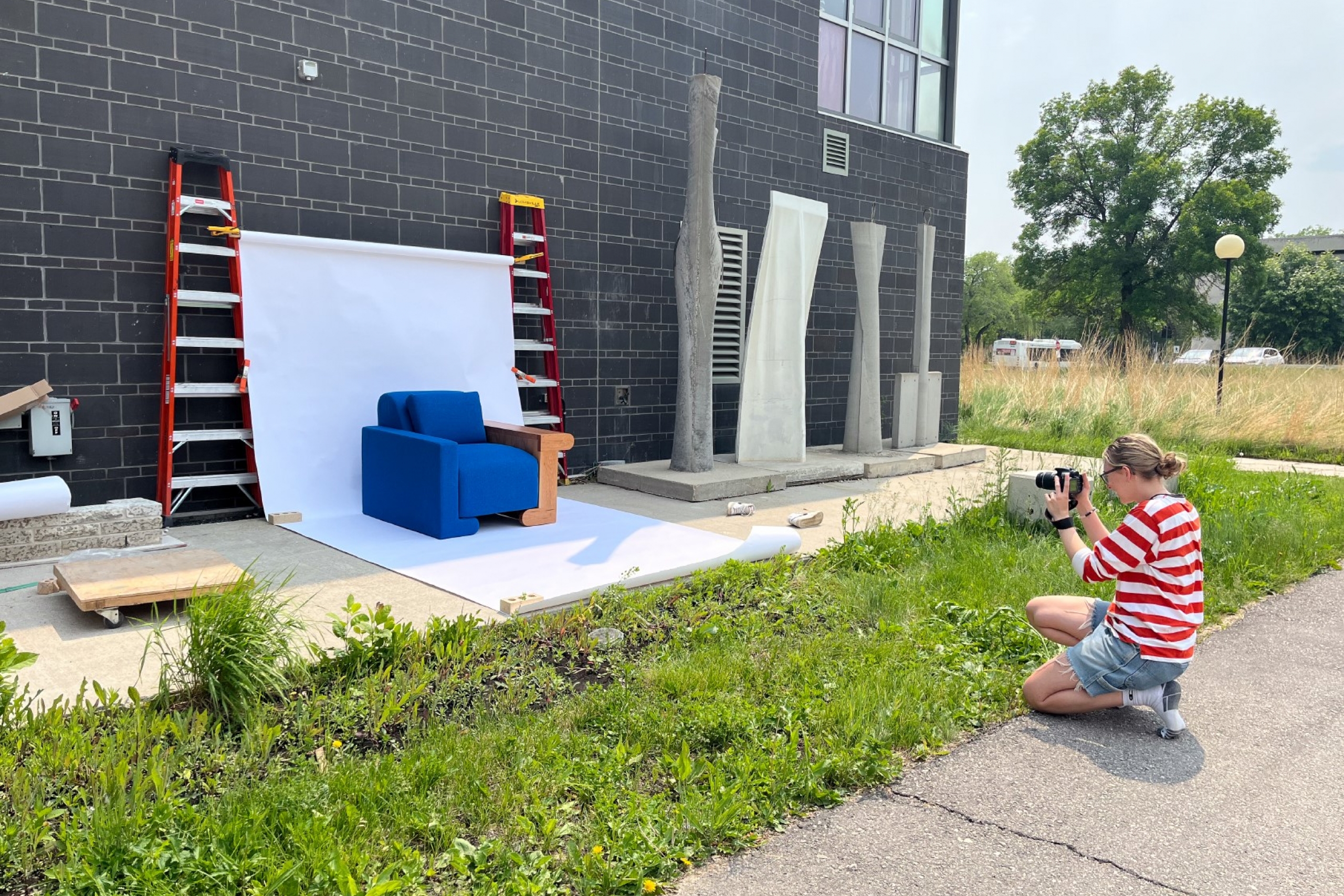 A person in a red-striped shirt takes a photo of a blue chair on a white backdrop outdoors, set against a dark brick wall. Sunny day, creative setup.
