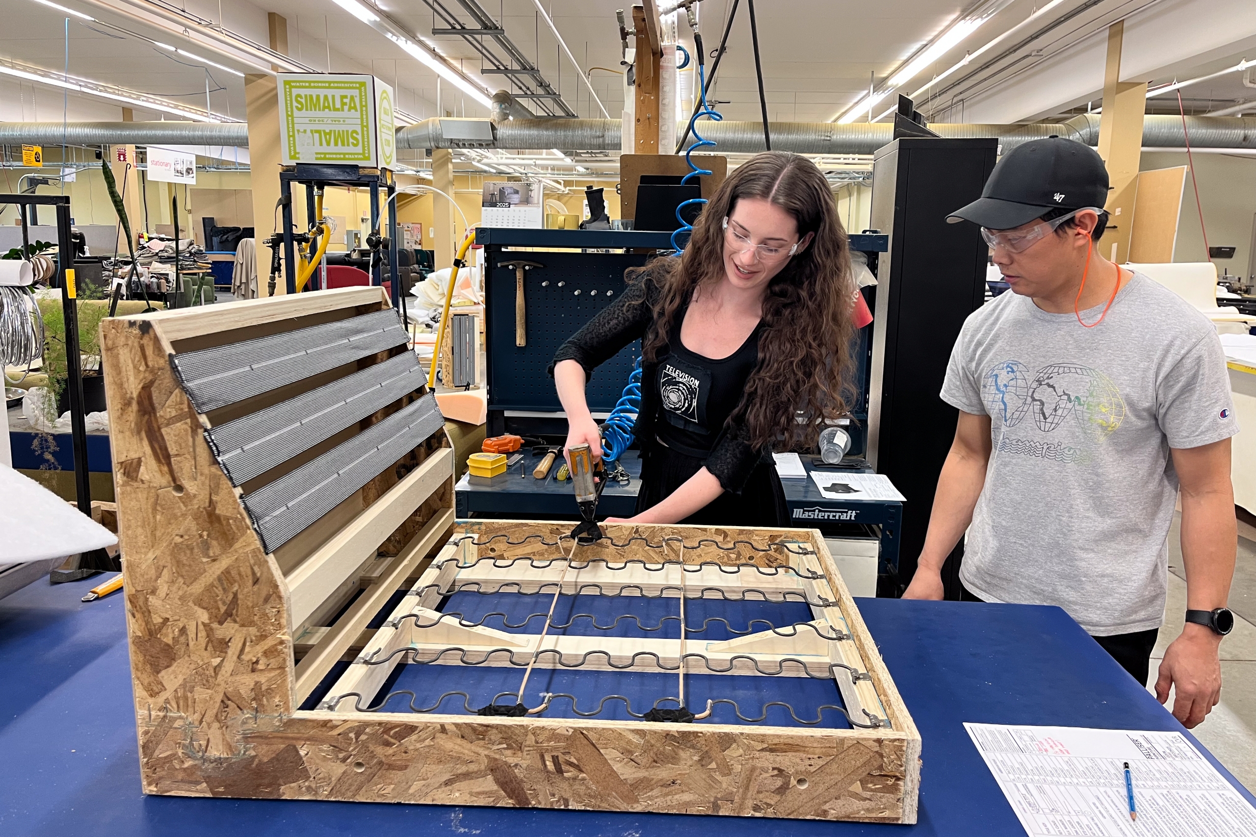 Two people work on a furniture frame in a workshop. The woman on the left uses a tool, while the man on the right observes. The setting is busy and industrious.