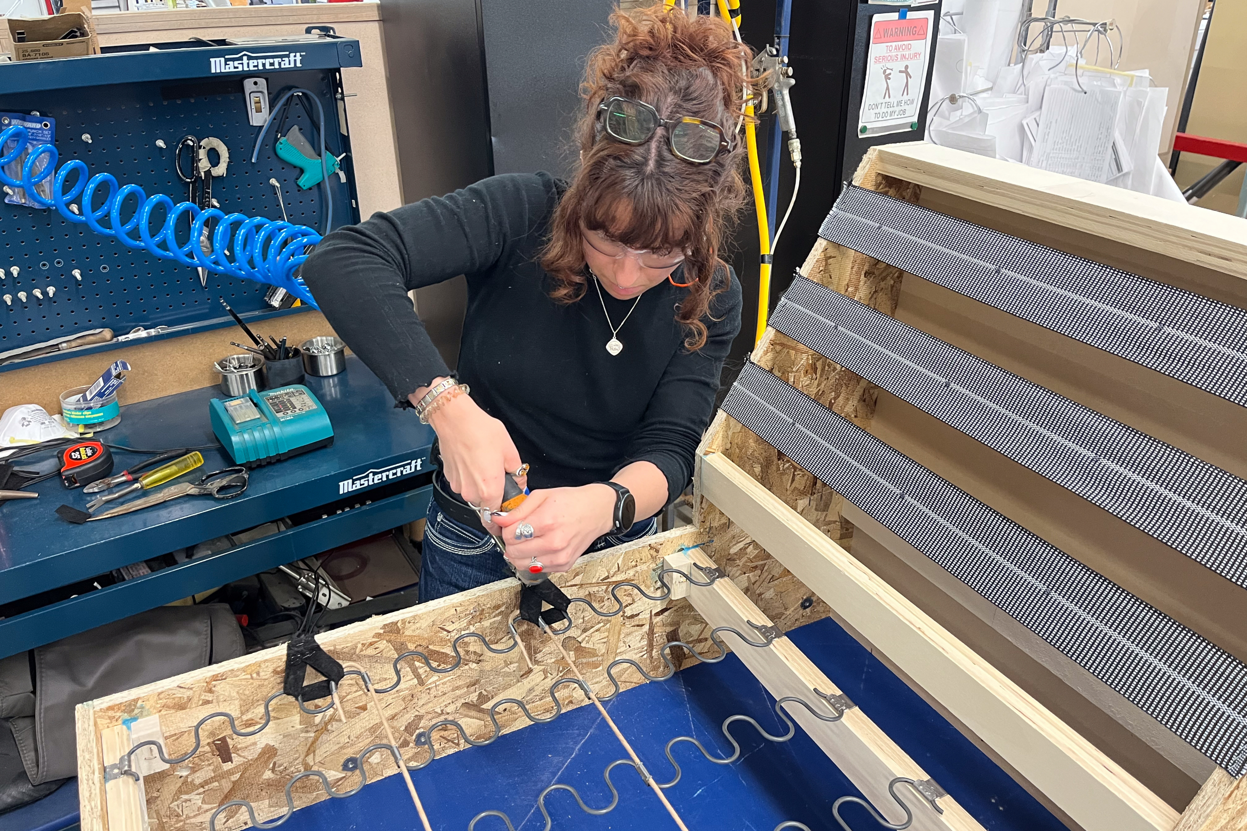 A woman in a workshop carefully works on a wooden frame with tools on the table. The scene conveys focus and craftsmanship in a DIY setting.