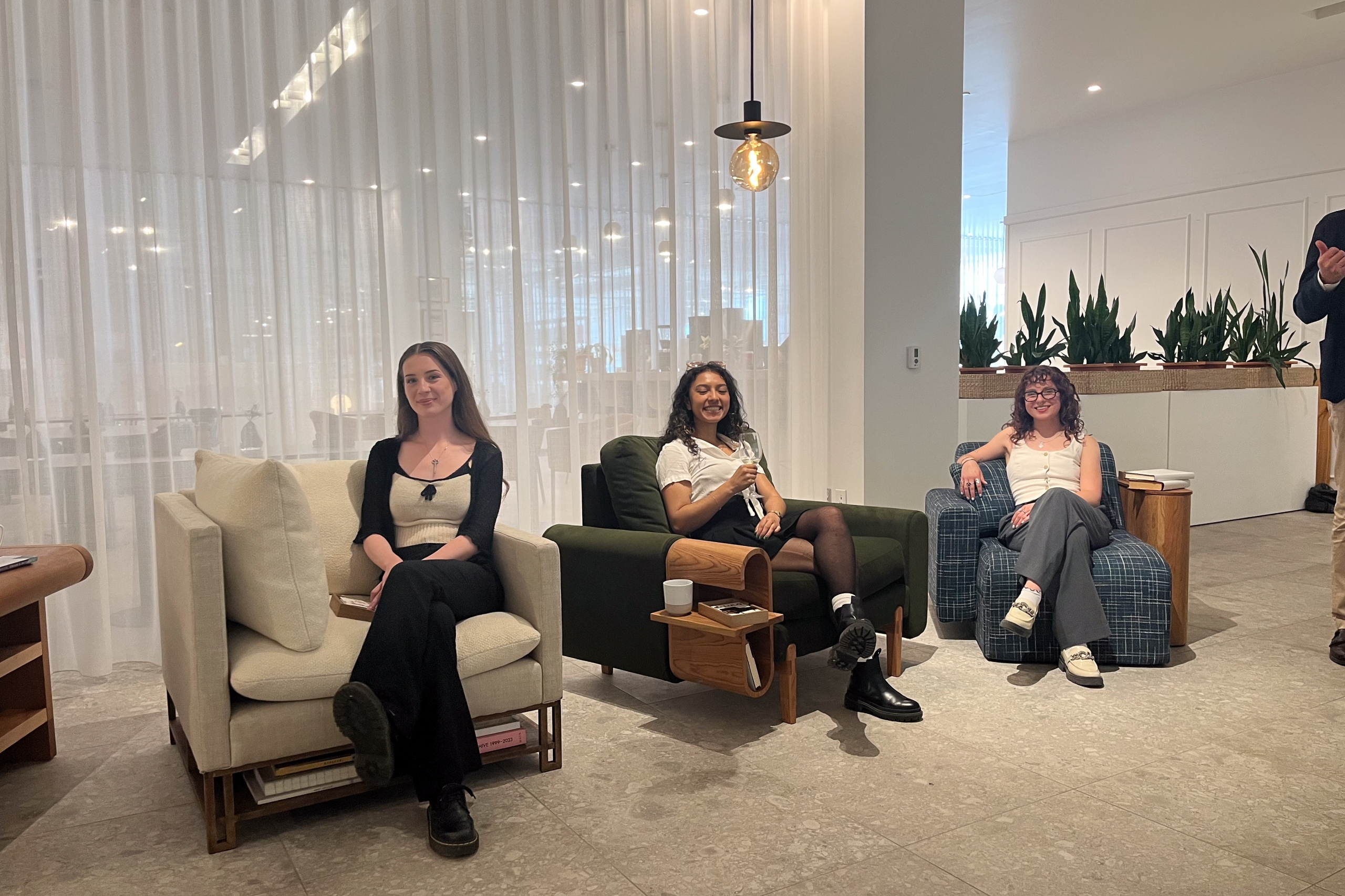 Three people smiling and sitting in comfortable chairs in a modern lounge with white curtains, plants, and warm lighting, conveying a relaxed atmosphere.