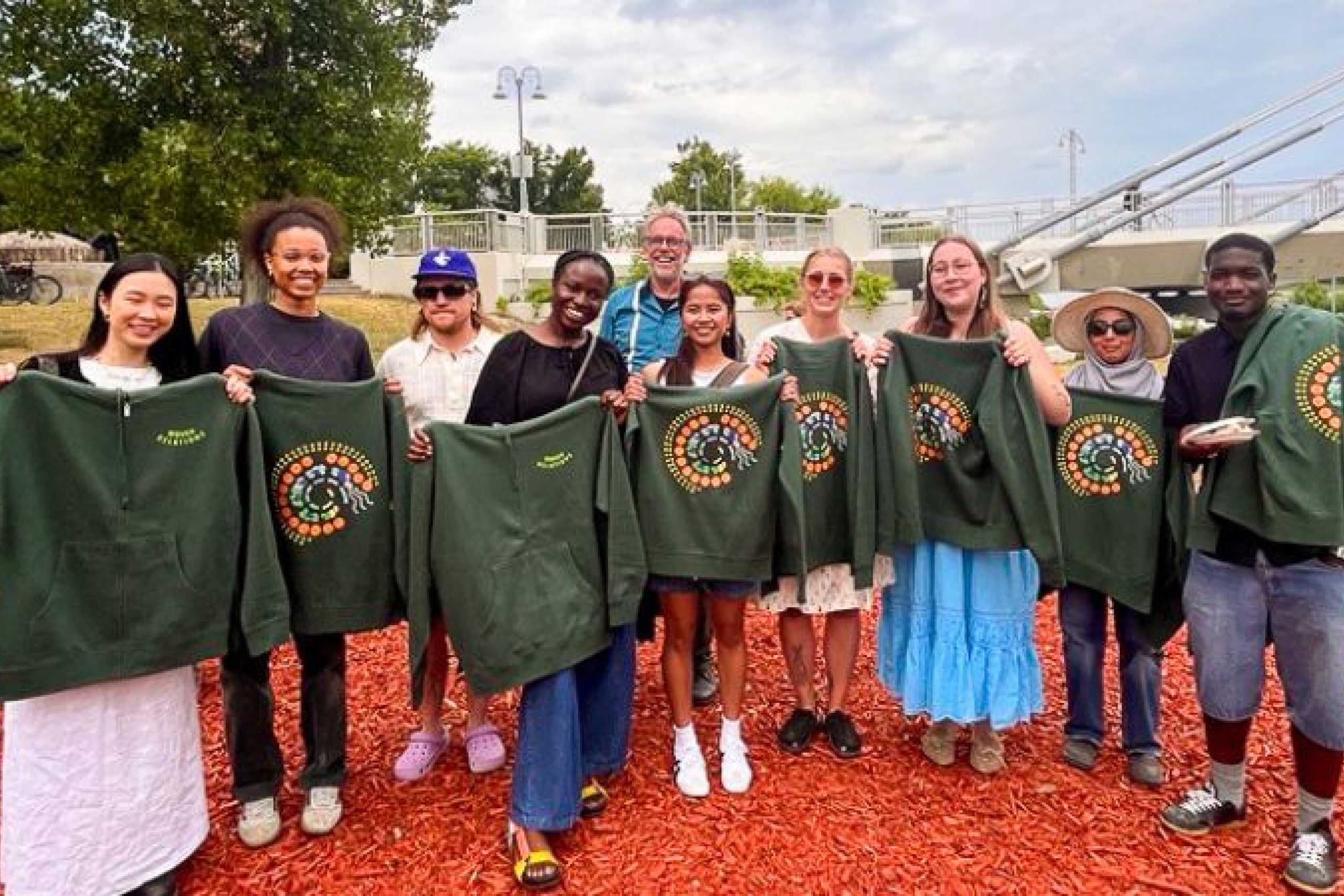 A diverse group of people smile outdoors, proudly holding green hoodies with colorful circular patterns. The mood is joyful and celebratory.