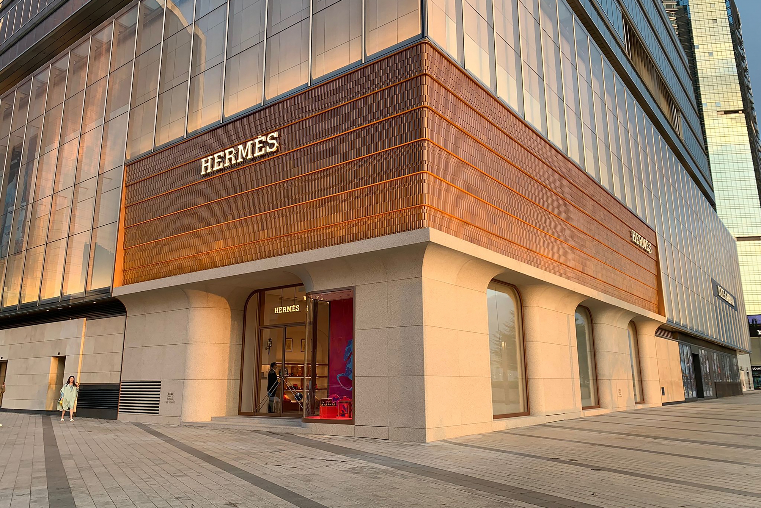 Corner view of the Hermès store with a modern glass and wood facade. Warm lighting creates an elegant atmosphere. A person walks nearby on the plaza.
