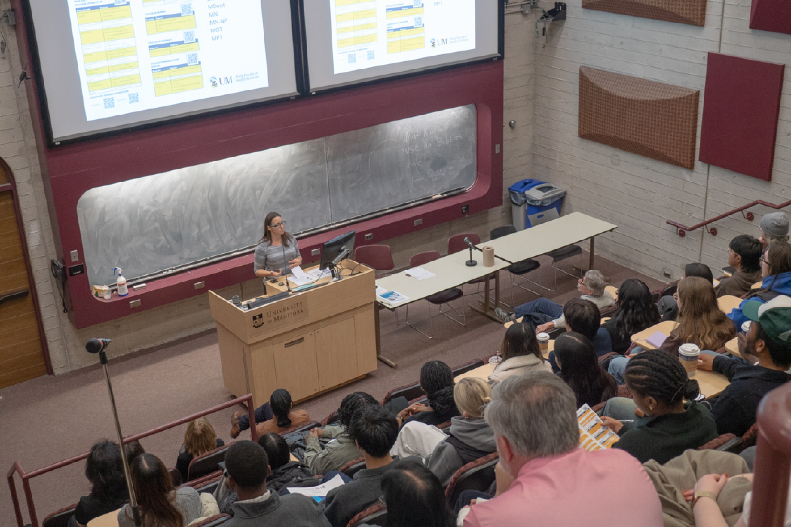 An amphitheatre style classroom is filled with students as they listen to a speaker.