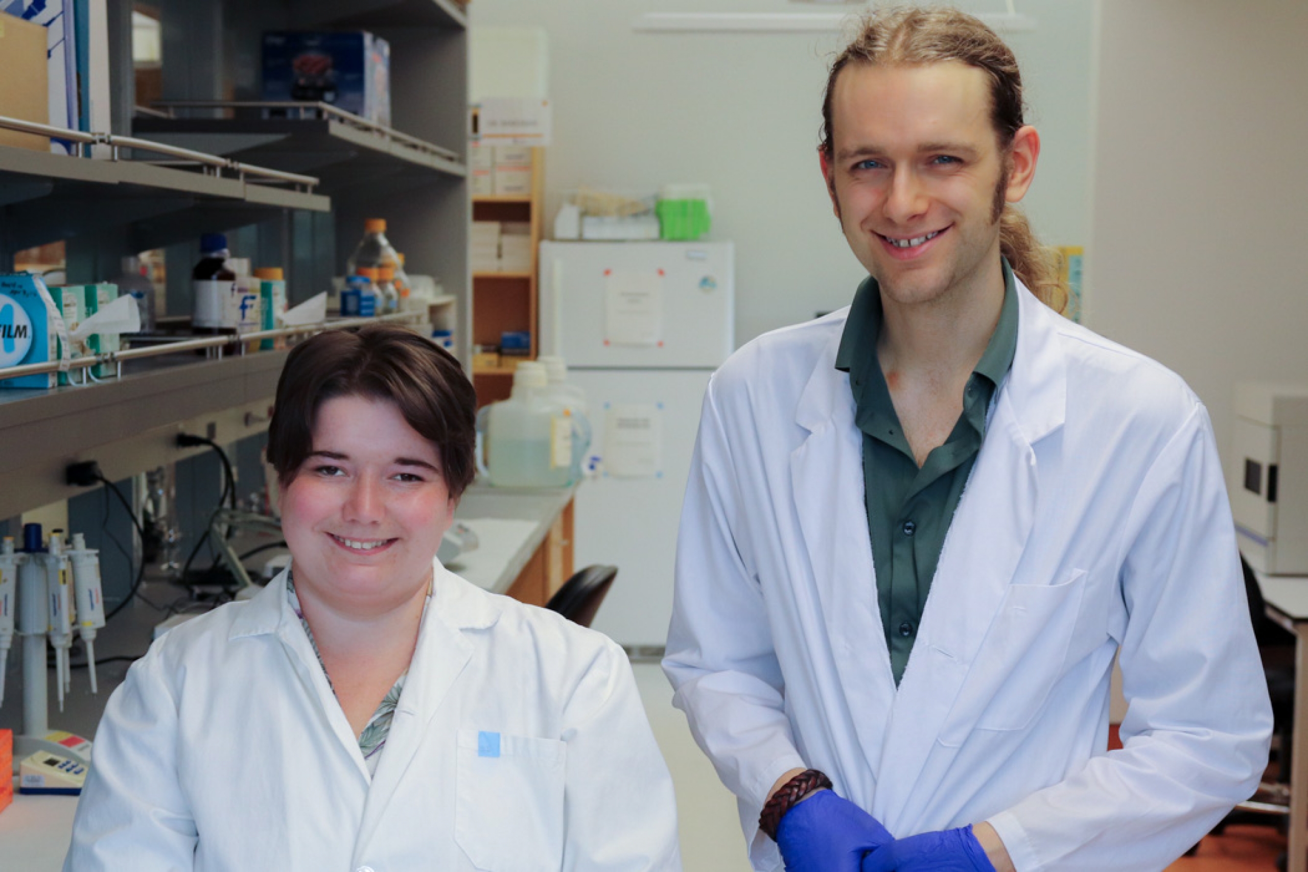 Janessa Sawatzky and Eli Mondor standing side by side in a science lab, smiling at the camera. Lab equipment and shelves are visible in the background.