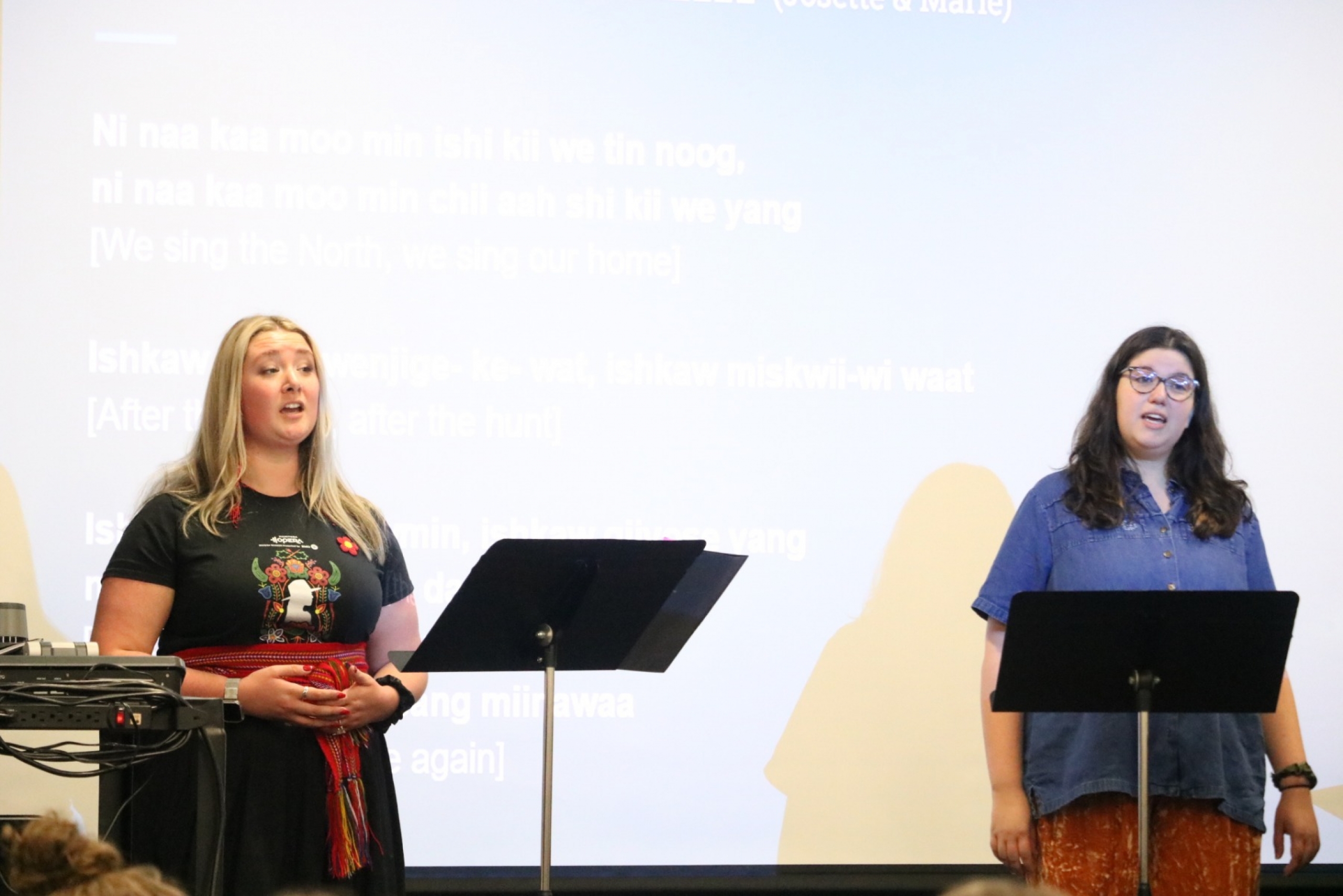 Two singing women stand in front of music podiums.