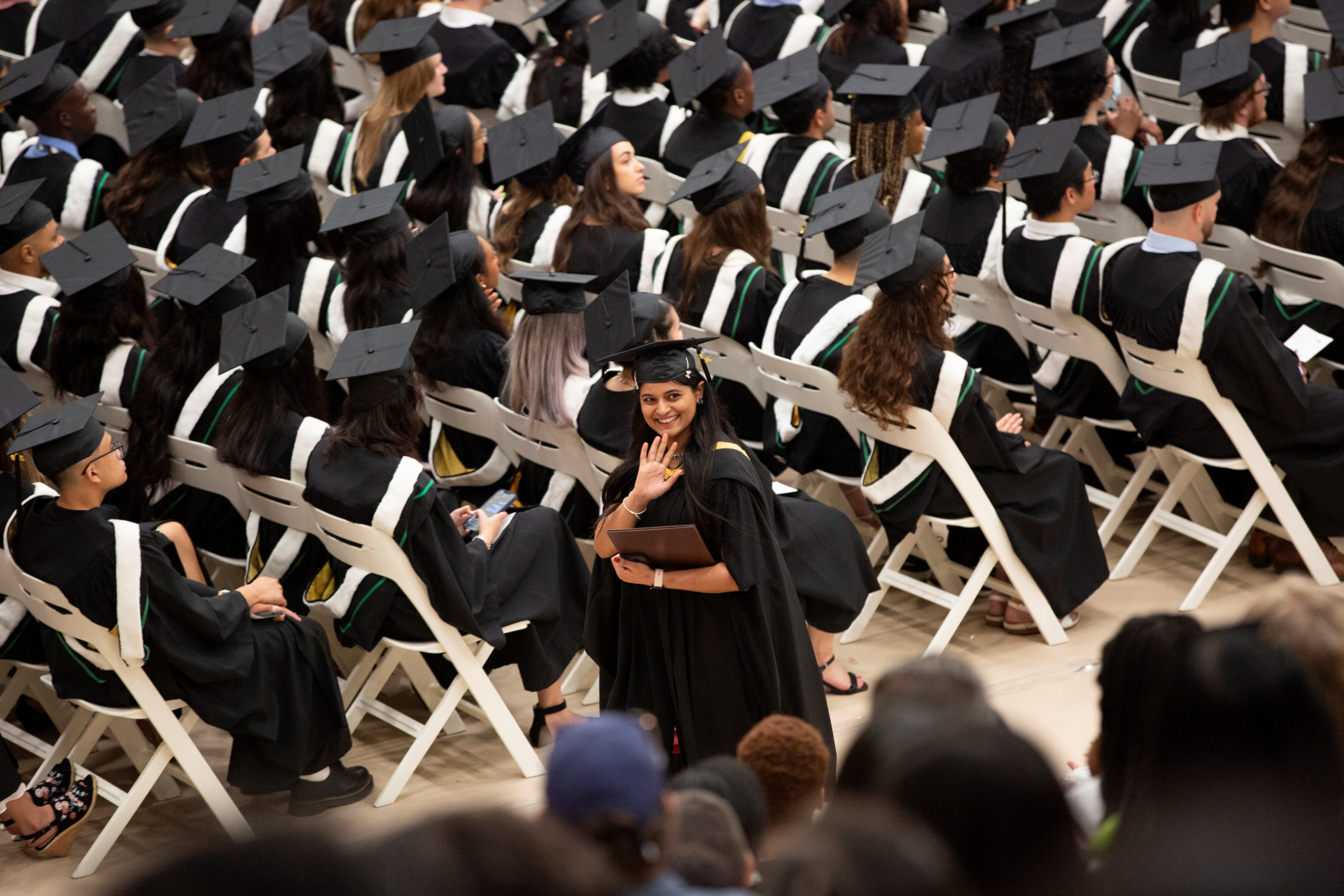 A graduating student smiles and waves in a sea of students wearing caps and gowns.