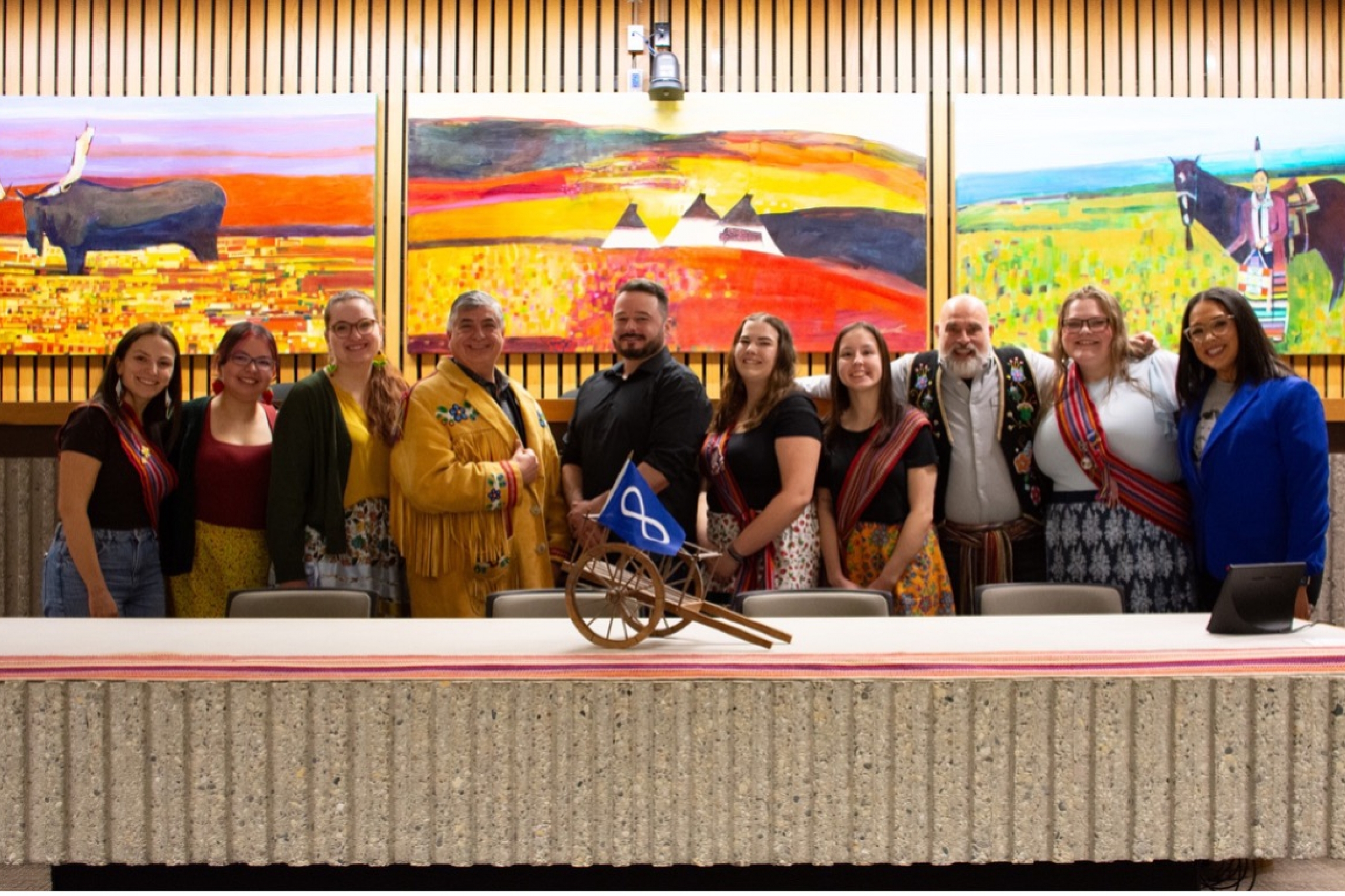 The panelists standing in front of colourful paintings at the Louis Riel Day event.
